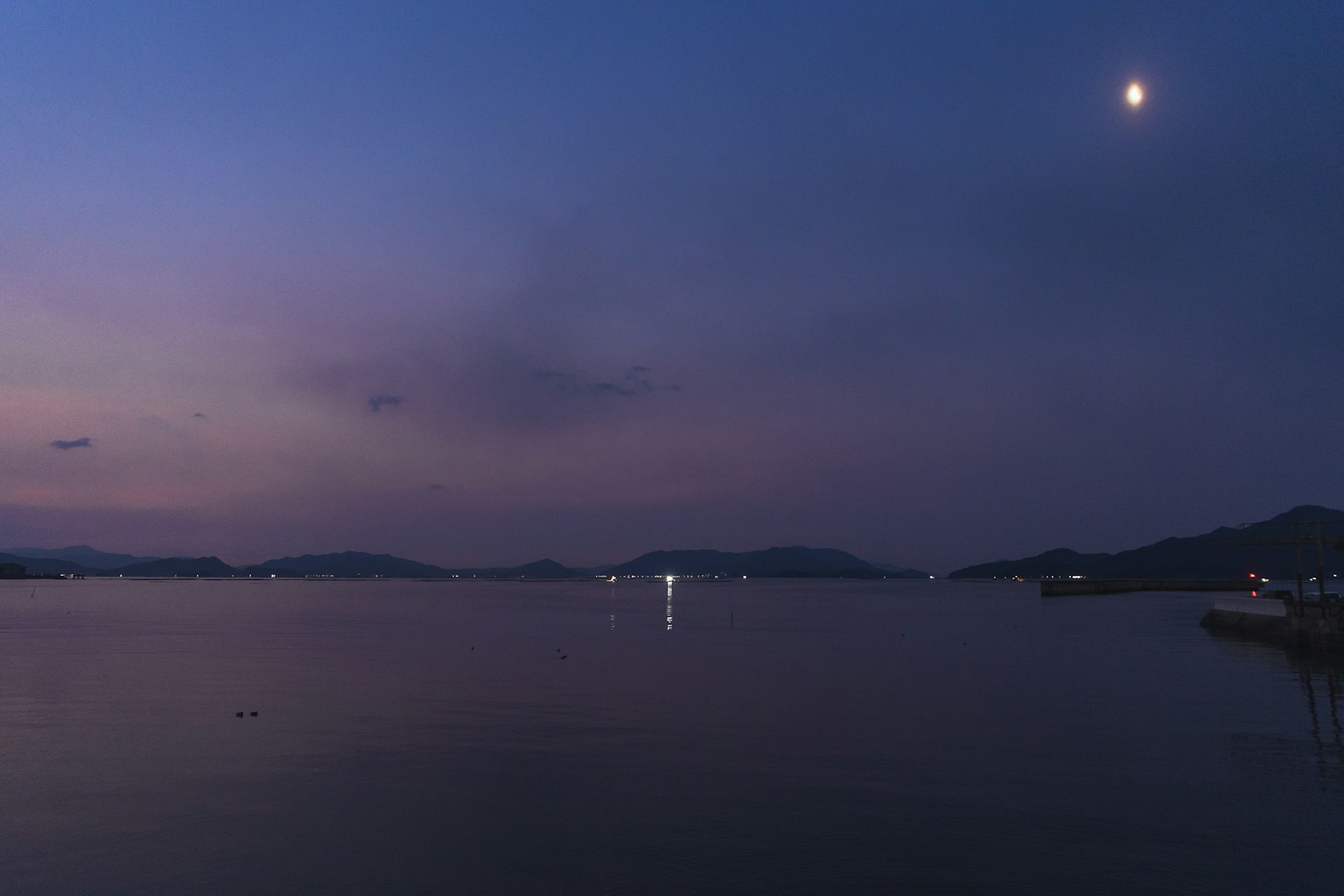 Ruhiges Meer in der Dämmerung mit Mondlicht und fernen Hügeln