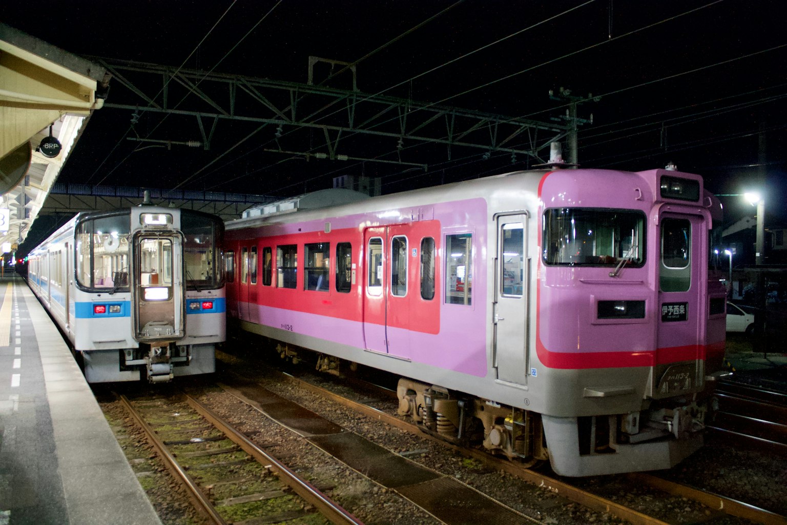 Two trains at a station during night time one pink and one gray