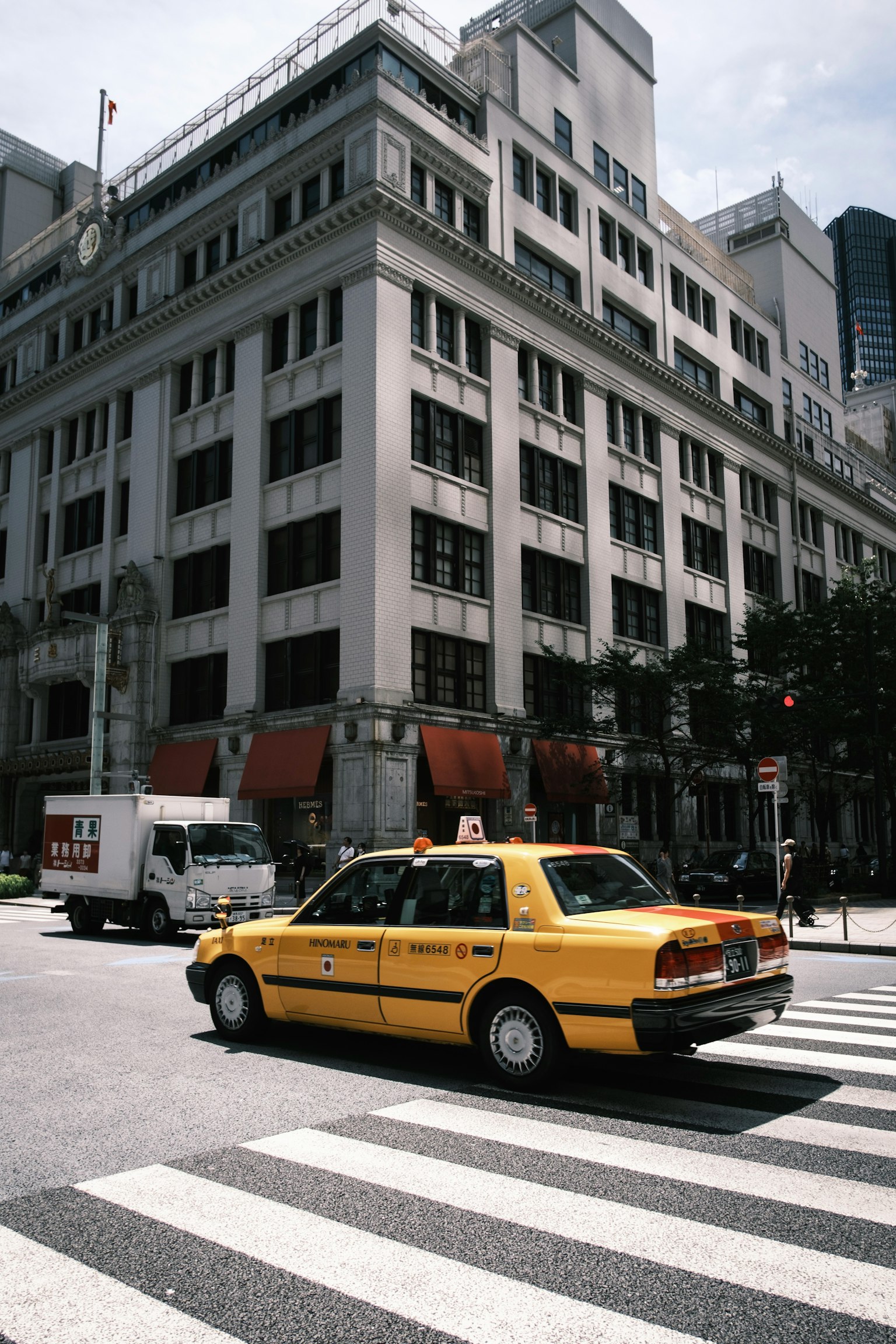 Một chiếc taxi màu vàng đang đi qua ngã tư trong bối cảnh đô thị với một tòa nhà cũ và kiến trúc hiện đại