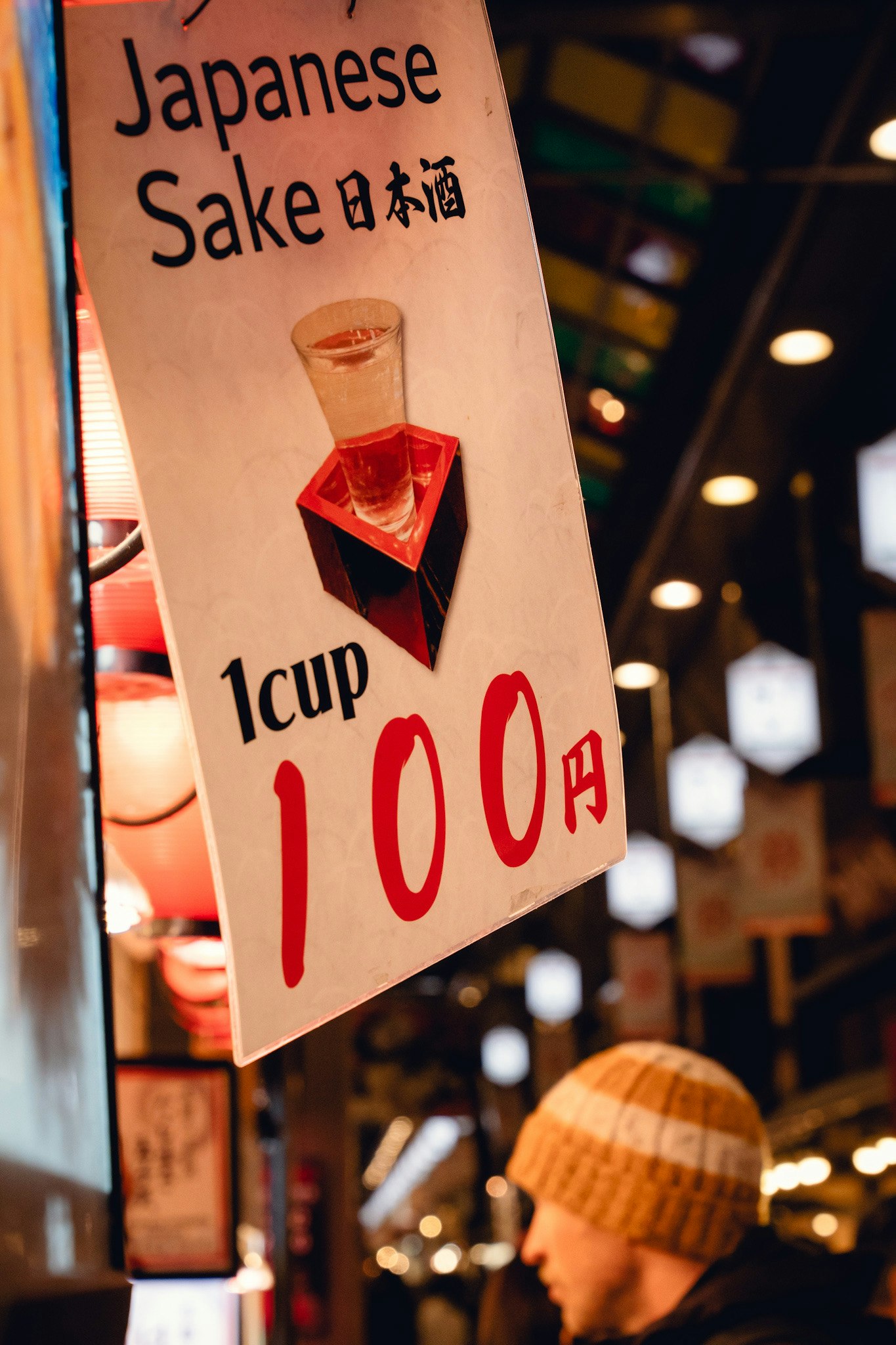 Suasana toko di dalam ruangan dengan papan bertuliskan “Japanese Sake 日本酒” dan “1 cup 100 yen”