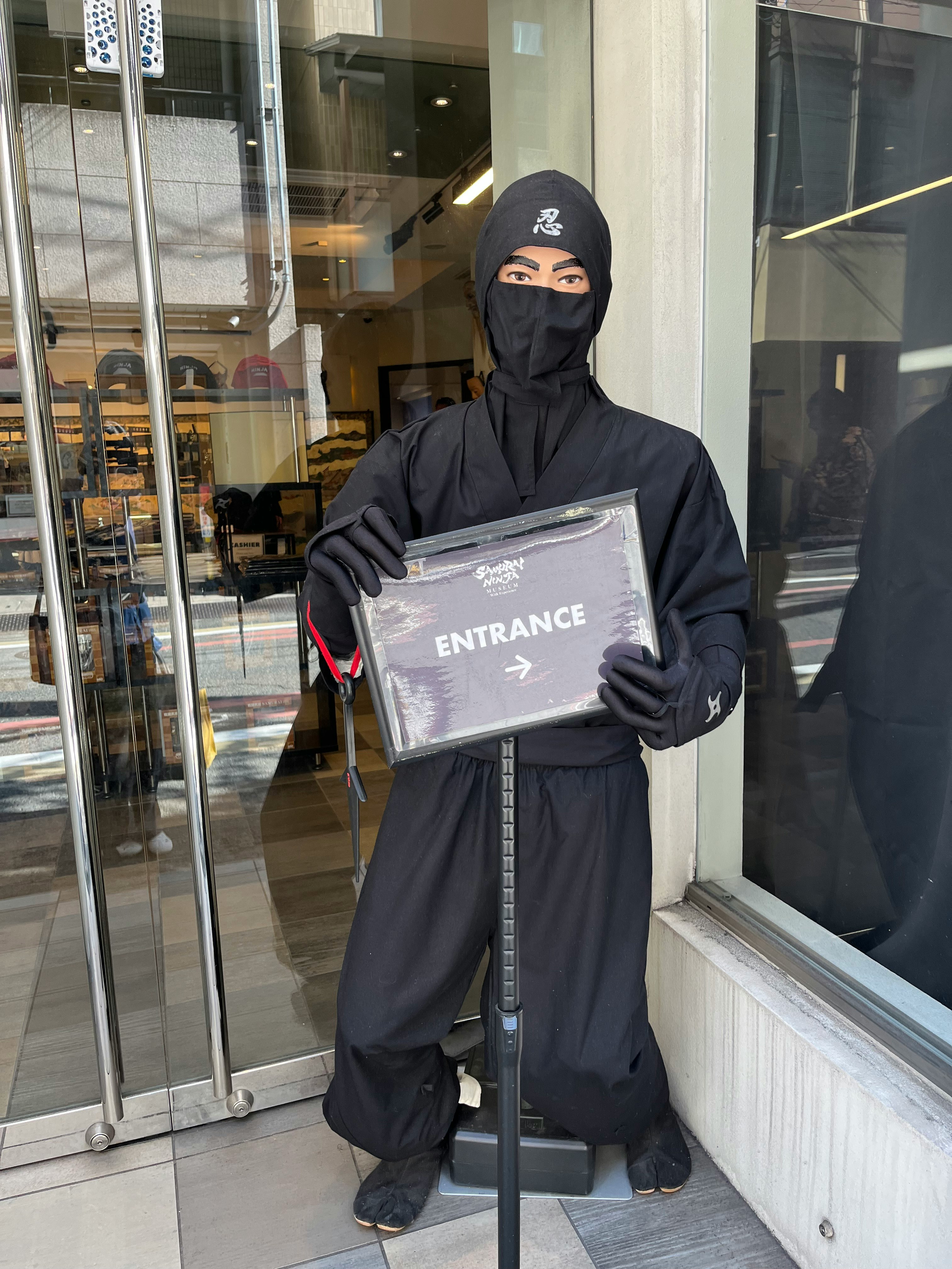 建物の入口前で「ENTRANCE」表示を持つ覆面の人物