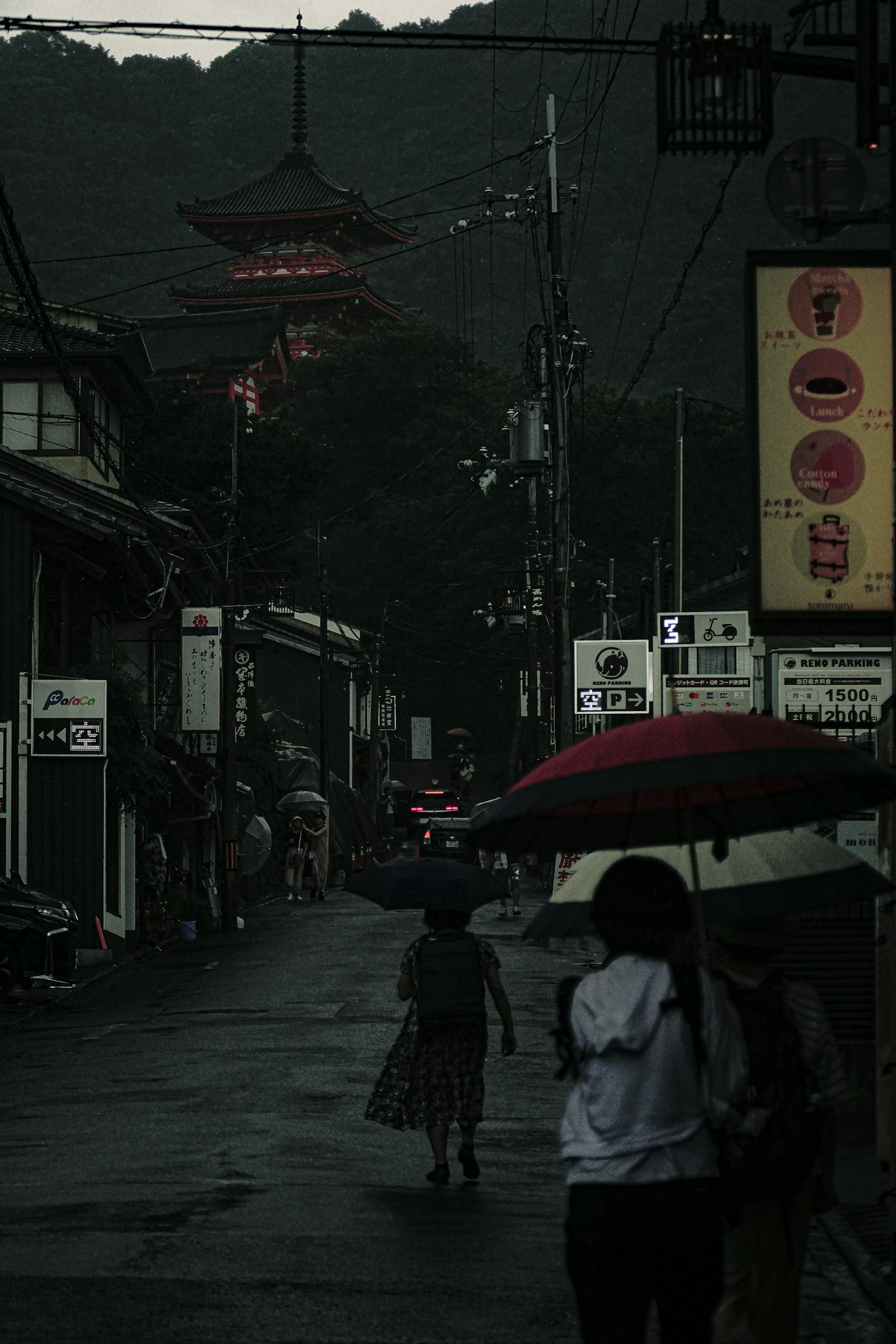 우산을 쓴 사람이 어두운 거리에서 걷고 있으며 배경에 산과 빨간 탑이 보인다