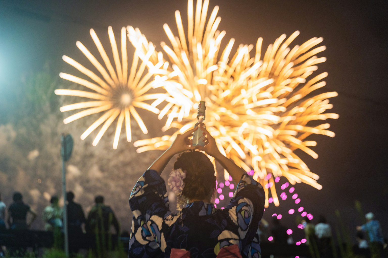 Một người trong trang phục yukata đang thưởng thức màn bắn pháo hoa với pháo hoa đầy màu sắc ở phía sau