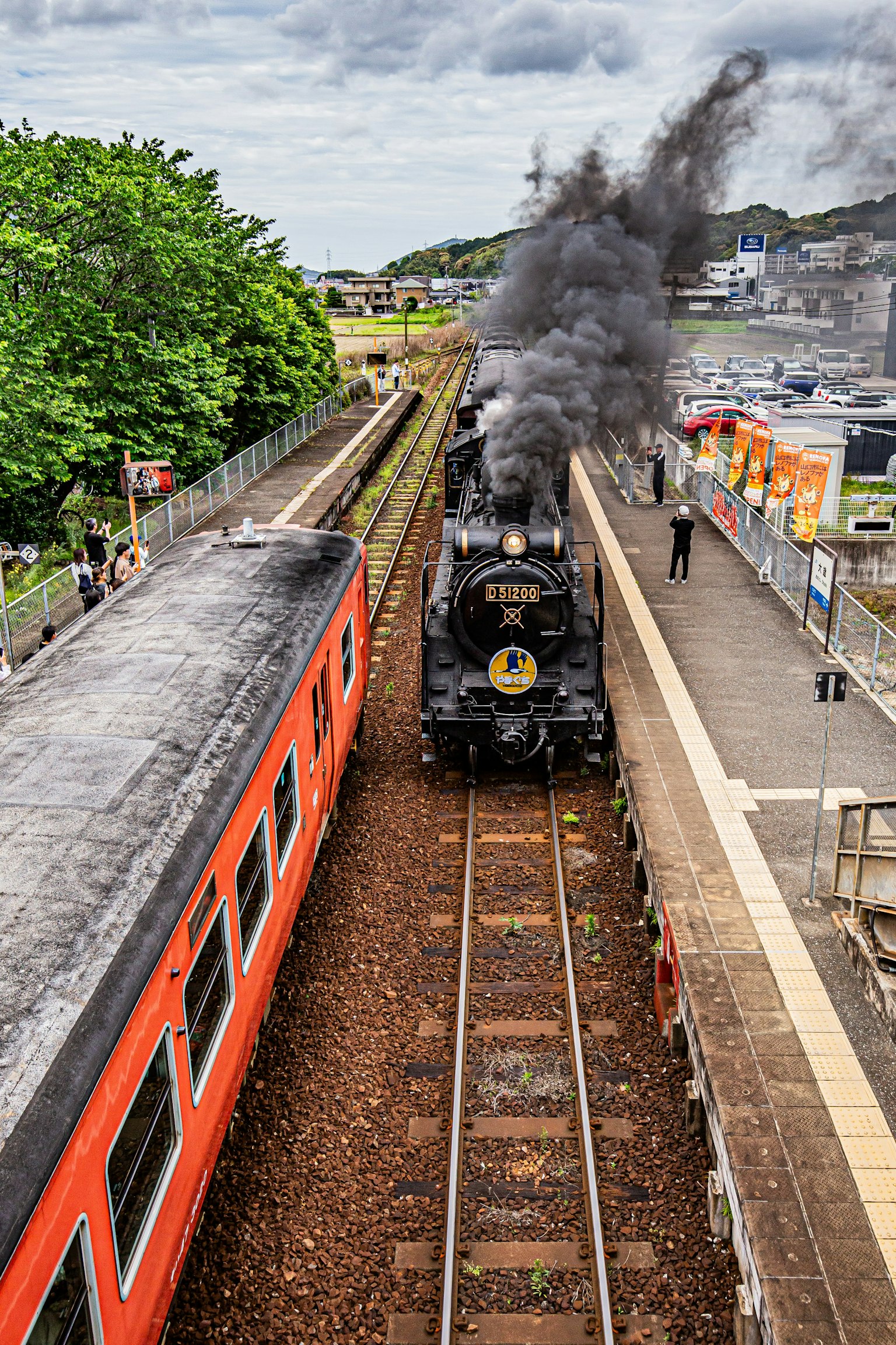Locomotora de vapor emitiendo humo con un tren rojo y una plataforma de estación visible