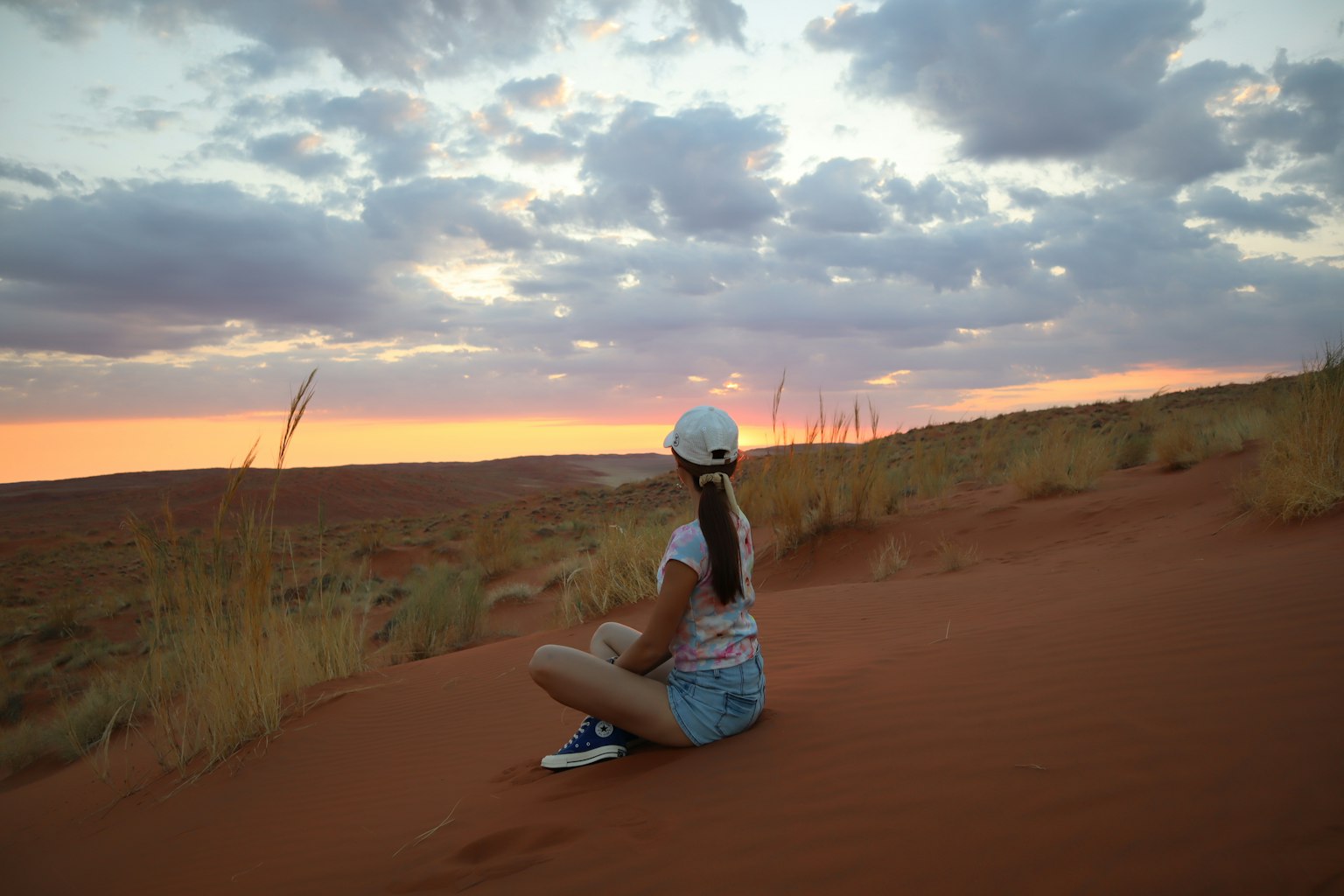 Fille assise sur le sable rouge regardant le coucher de soleil Détente dans la nature avec de l'herbe et des dunes