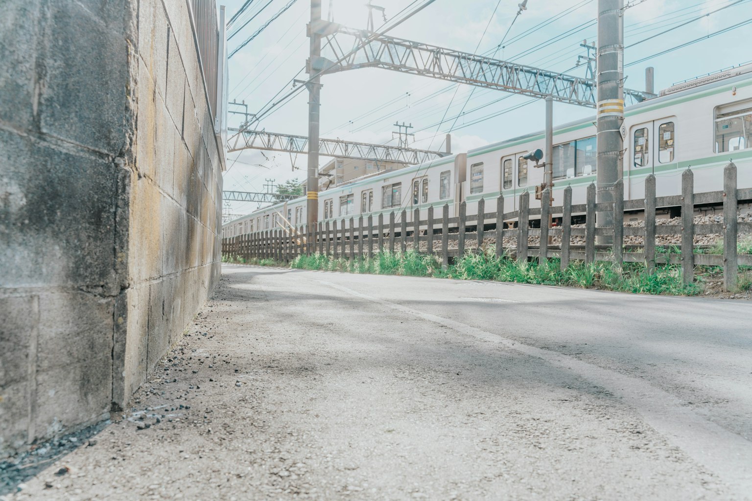 View of a railway side path with a train and overhead wires under a blue sky