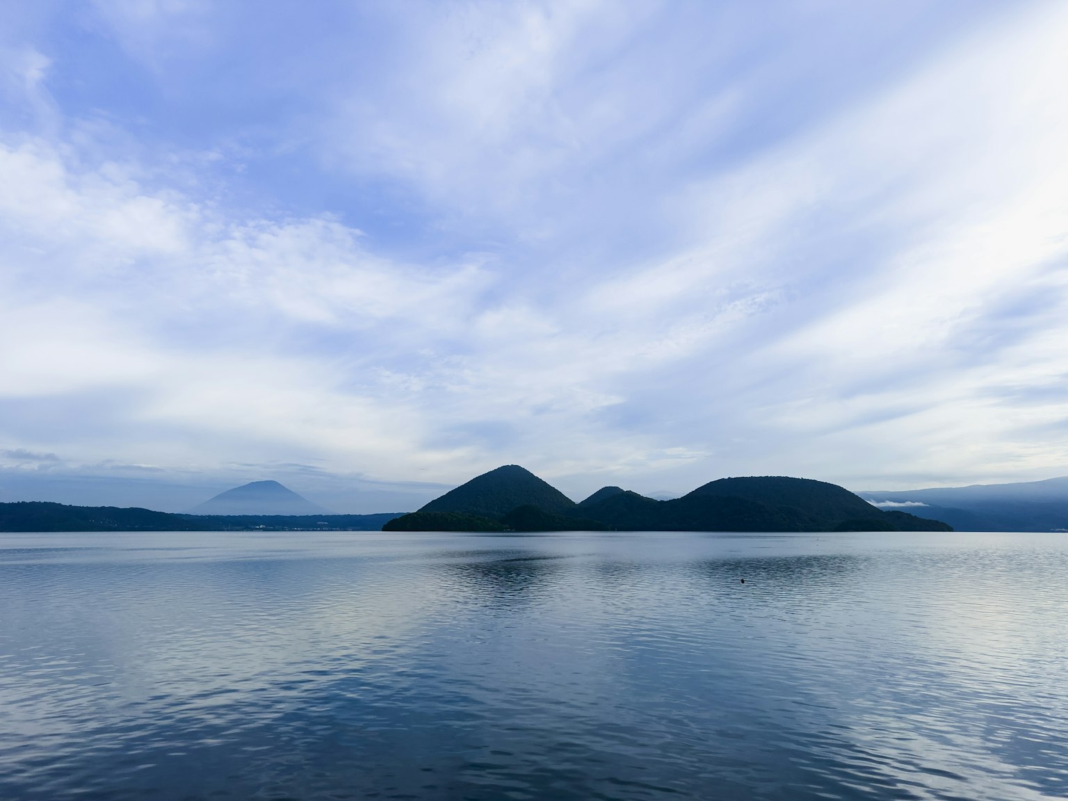Ruhiger See, der einen blauen Himmel und Wolken mit Silhouetten von Bergen spiegelt