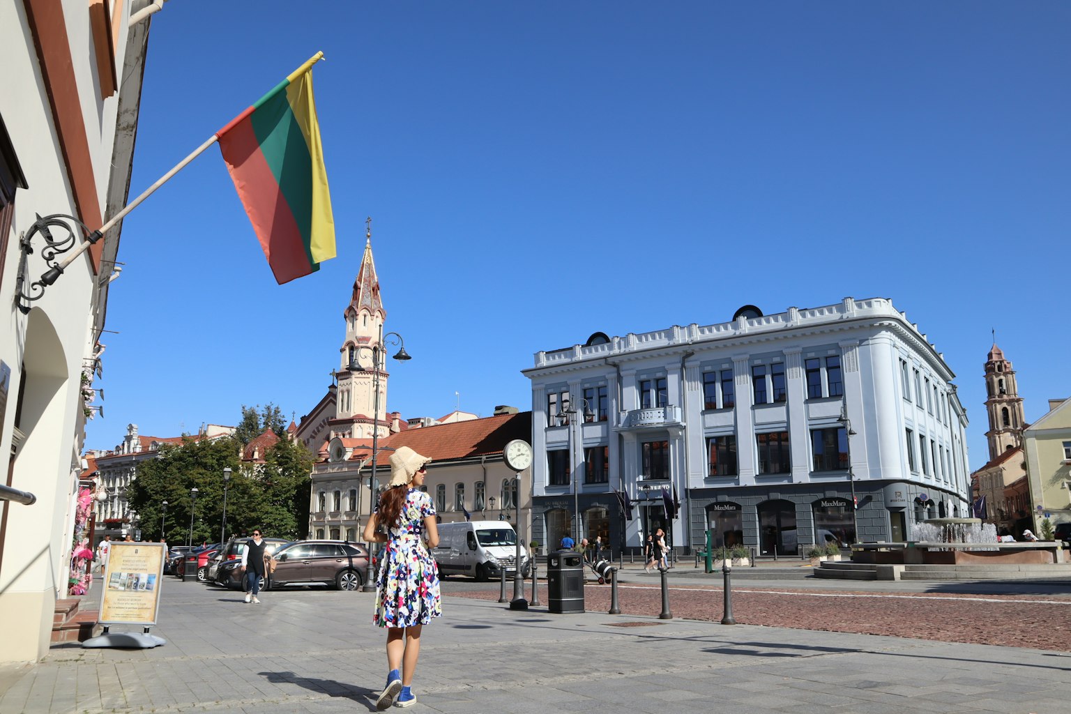 A person walking in a street scene with the Lithuanian flag under a clear blue sky