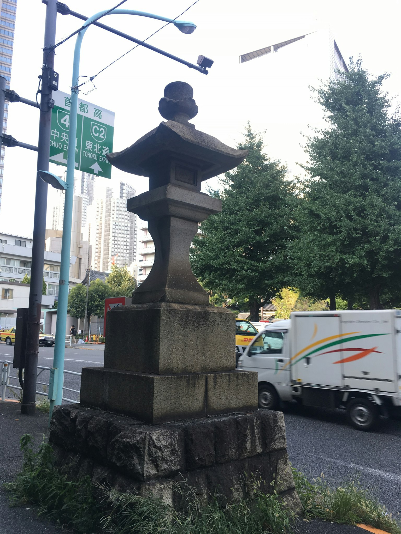 Una antigua linterna de piedra en una esquina de la calle rodeada de árboles verdes y vehículos