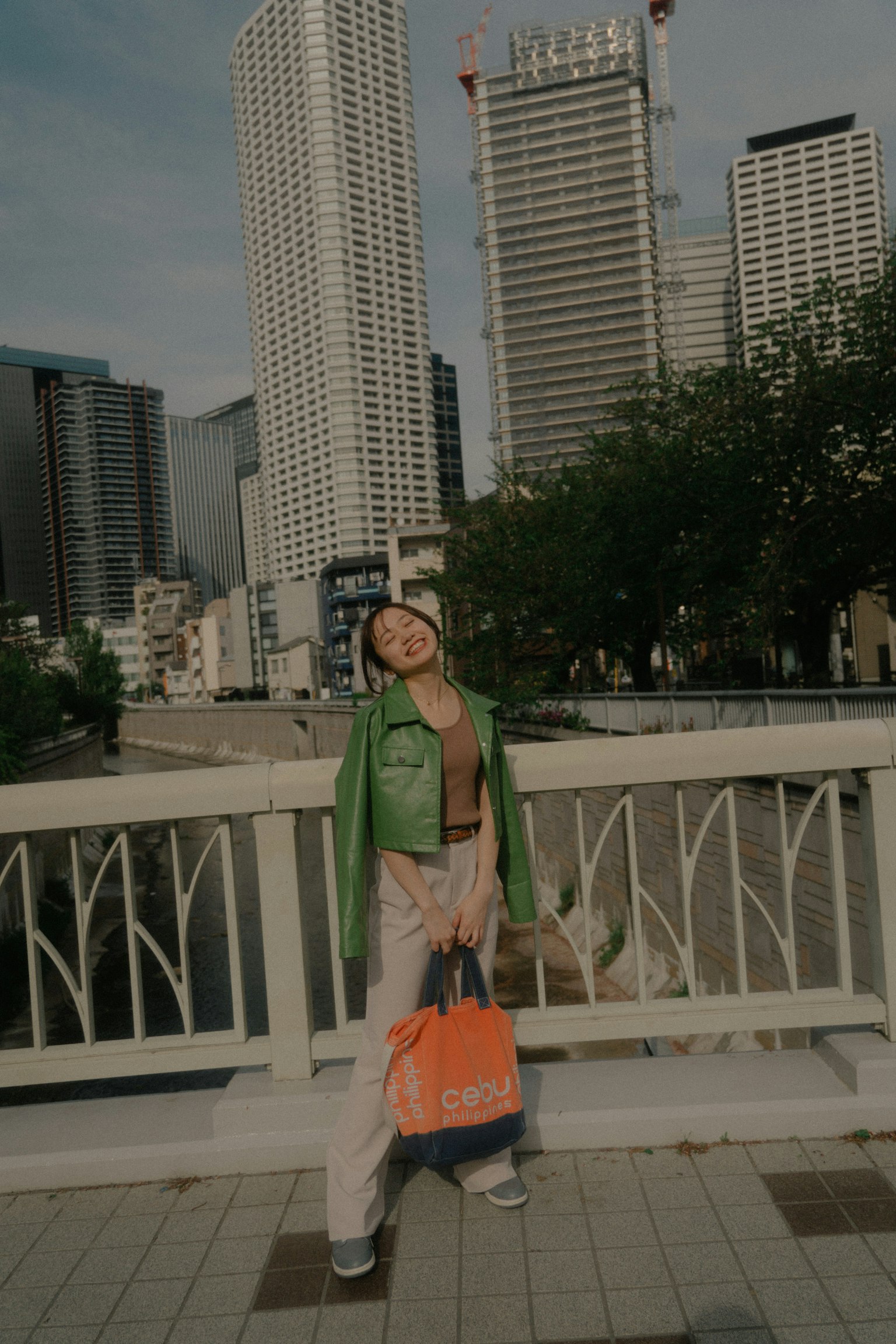 Fashion model posing in urban landscape wearing a green jacket and holding an orange bag