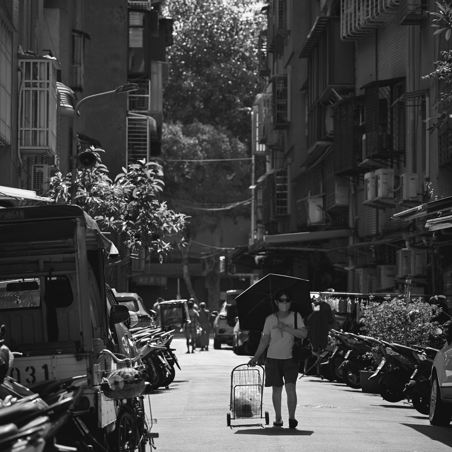 Una persona che cammina in un vicolo stretto circondato da motociclette e tricicli in un paesaggio urbano in bianco e nero