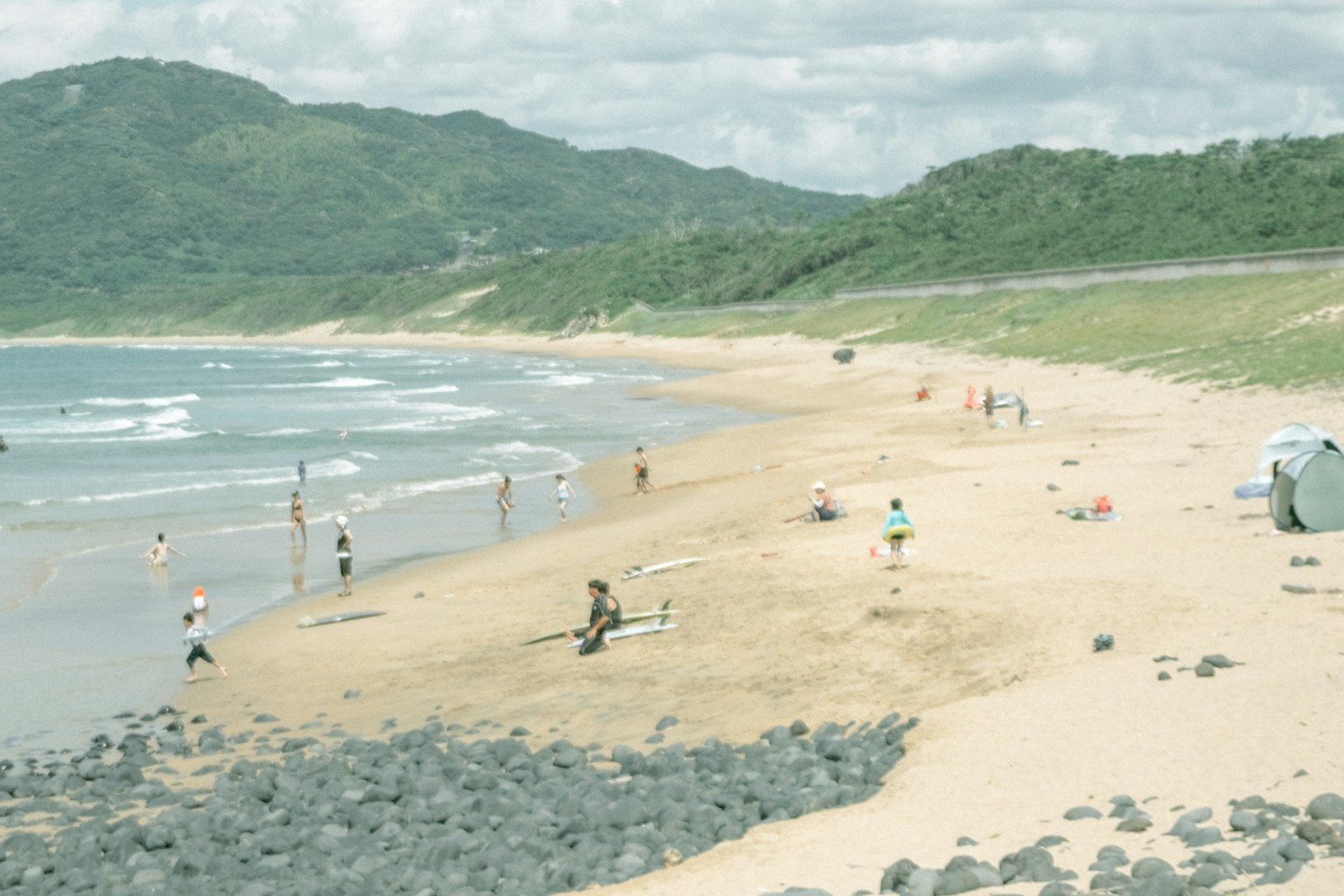 Pemandangan pantai dengan orang-orang menikmati pasir dan ombak dikelilingi bukit hijau