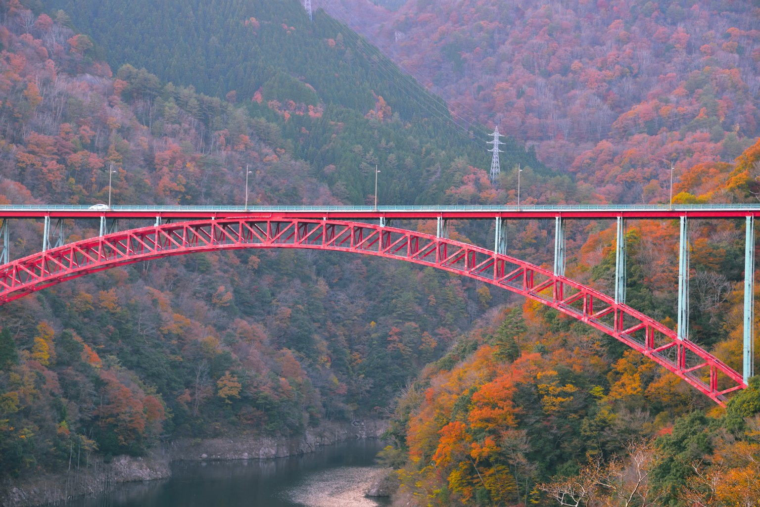 สะพานโค้งสีแดงล้อมรอบด้วยต้นไม้ฤดูใบไม้ร่วงที่มีสีสันและแม่น้ำ