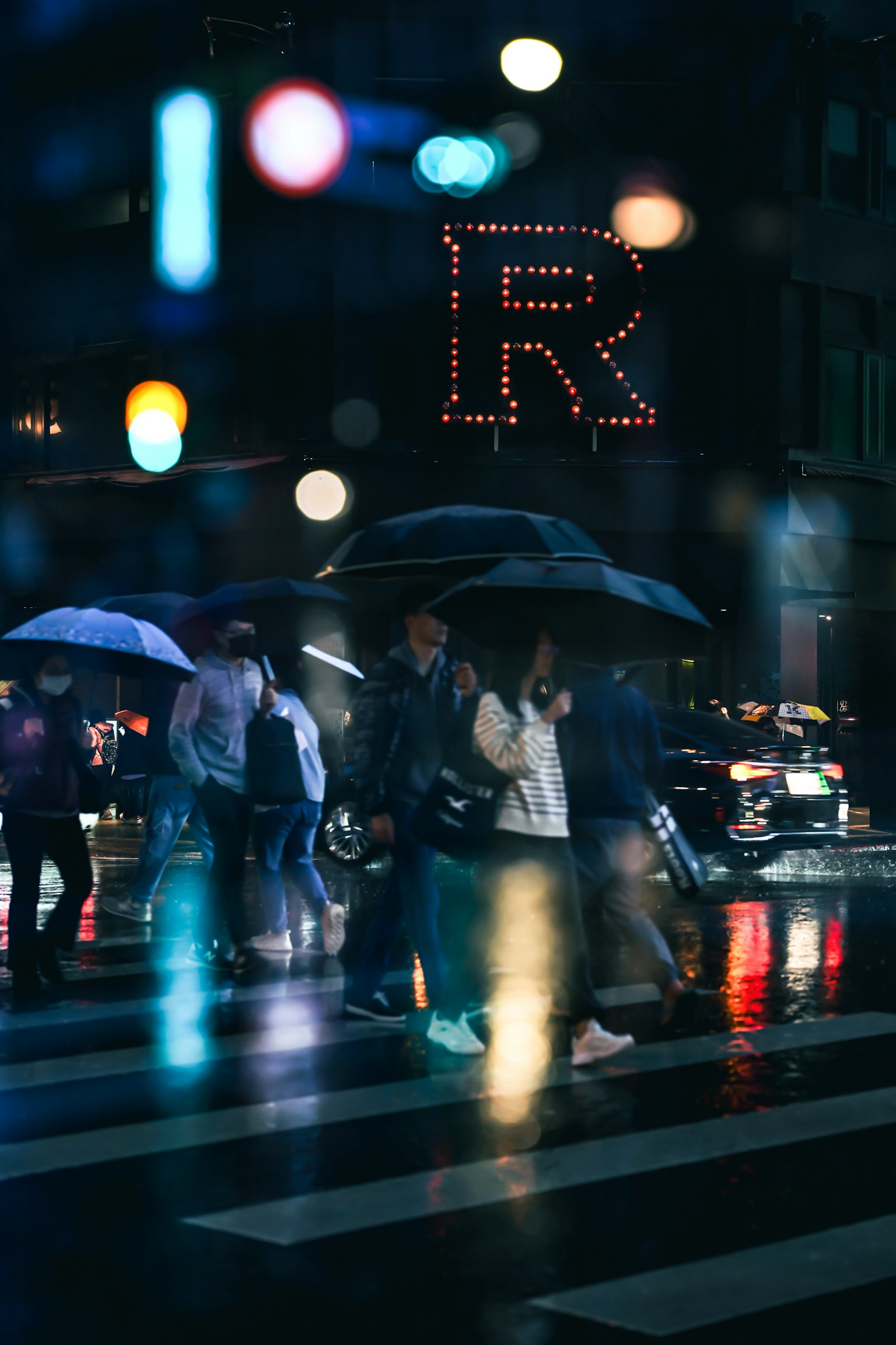 Người cầm ô băng qua đường trong mưa vào ban đêm với biển quảng cáo R neon ở phía sau