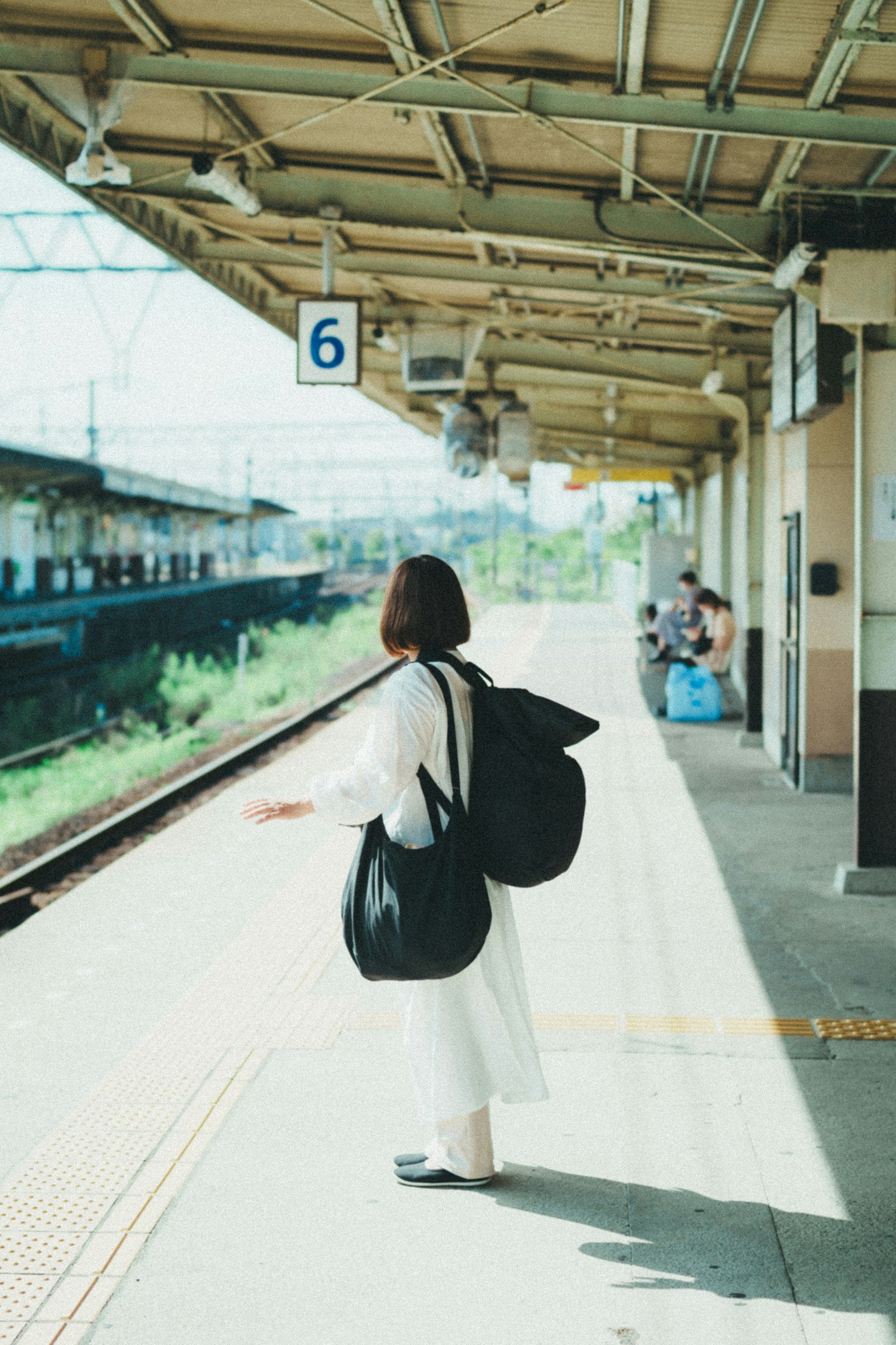 A woman standing on a train platform wearing white clothing and carrying a large bag