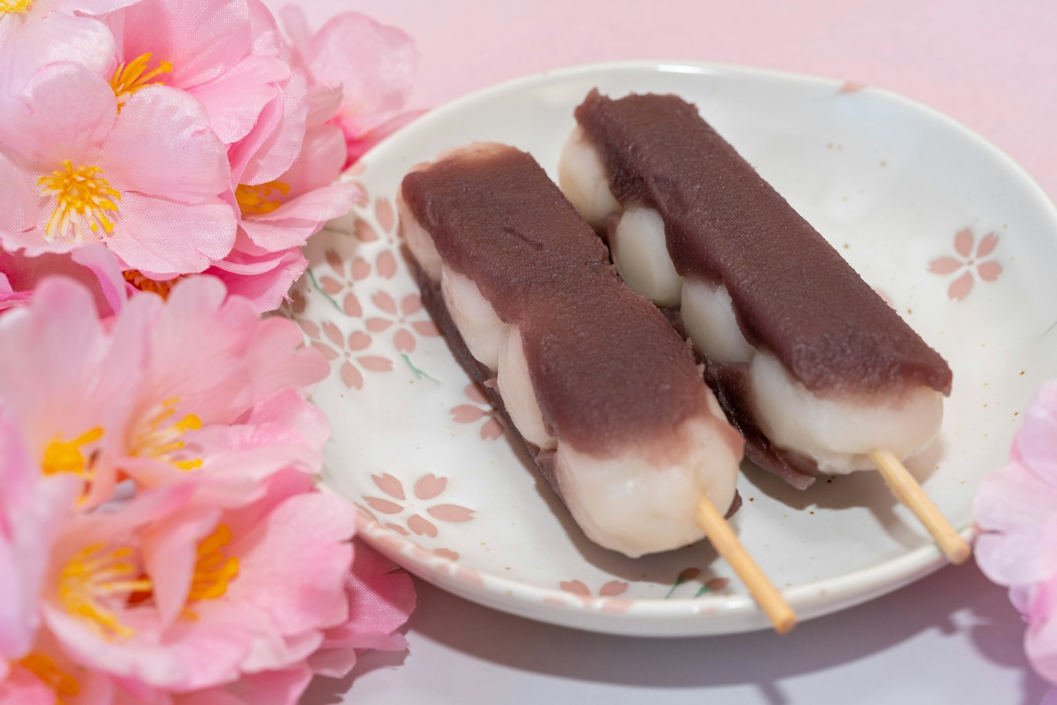 Due spiedini di dango con pasta di fagioli rossi su un piatto floreale circondato da fiori di ciliegio