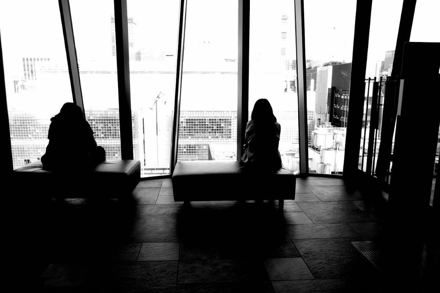 Siluetas de dos personas sentadas cerca de grandes ventanas en un entorno monocromático