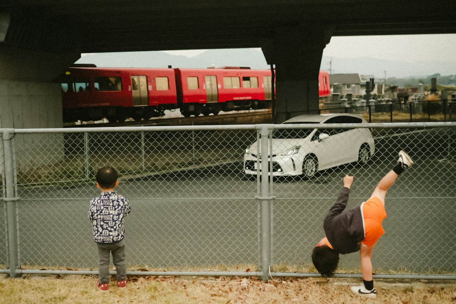 Children playing near a road with a red train and a white car