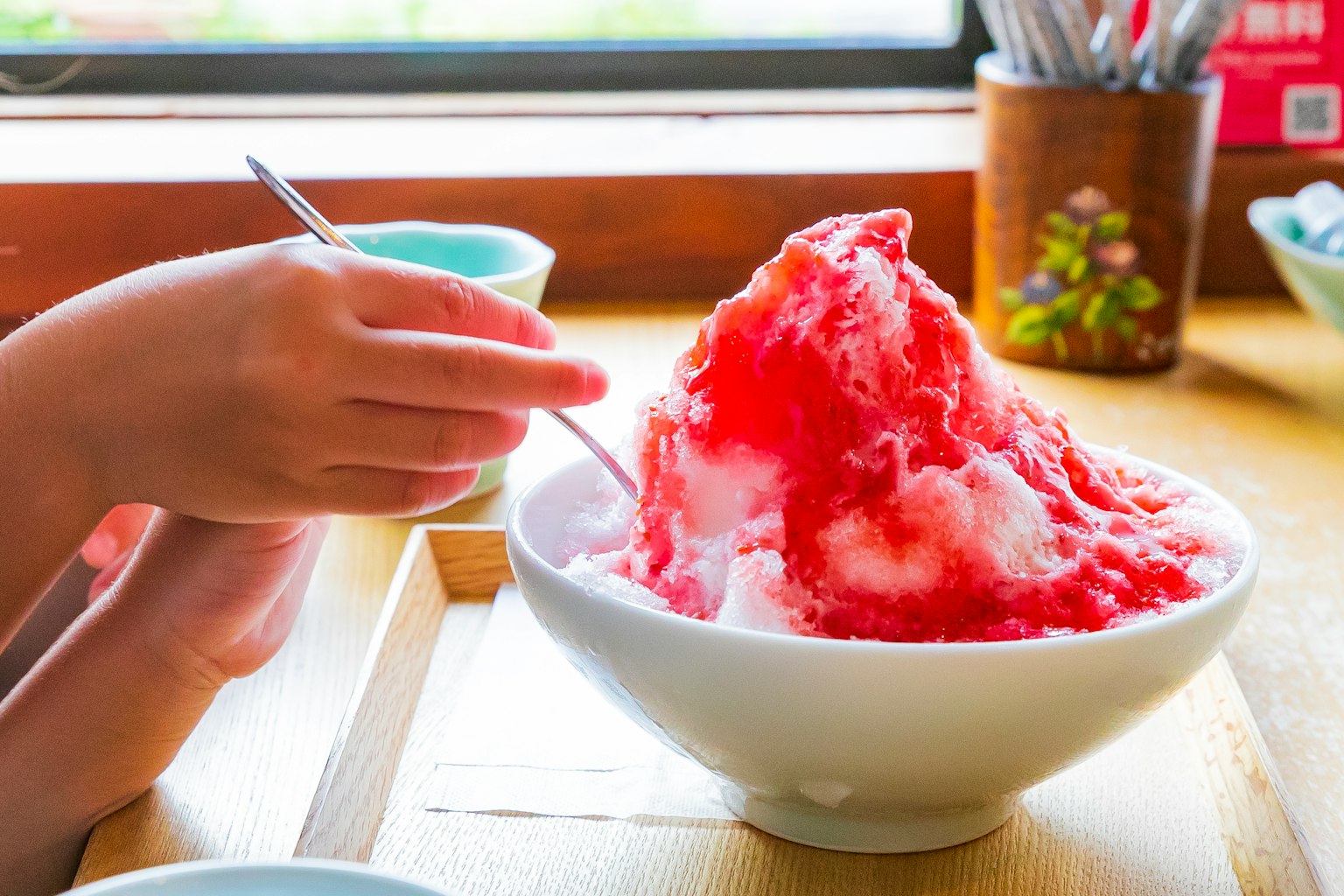 A hand holding a spoon over a large bowl of shaved ice topped with red syrup