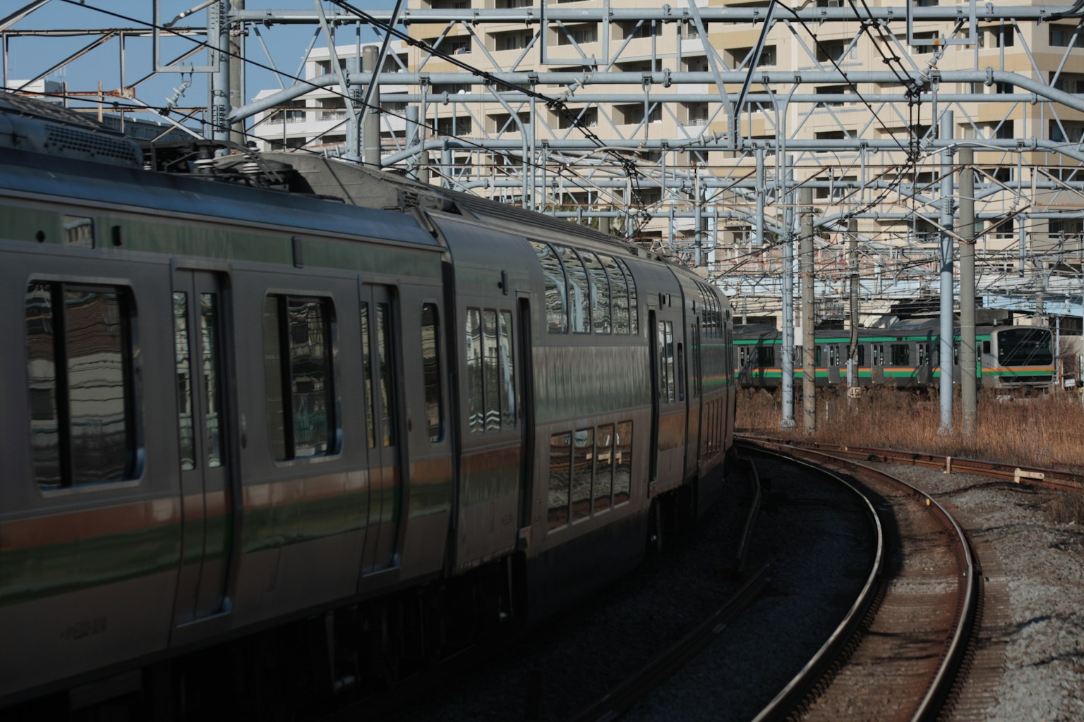 Treno argento che curva sui binari con un vecchio edificio sullo sfondo e linee aeree prominenti