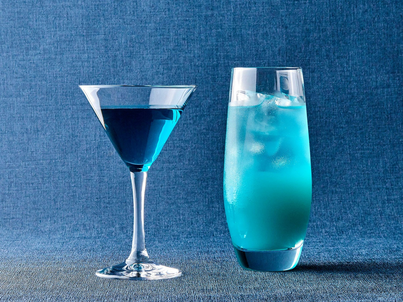 Zwei blaue Getränke ein Martiniglas und ein hohes Glas mit Eis vor blauem Hintergrund