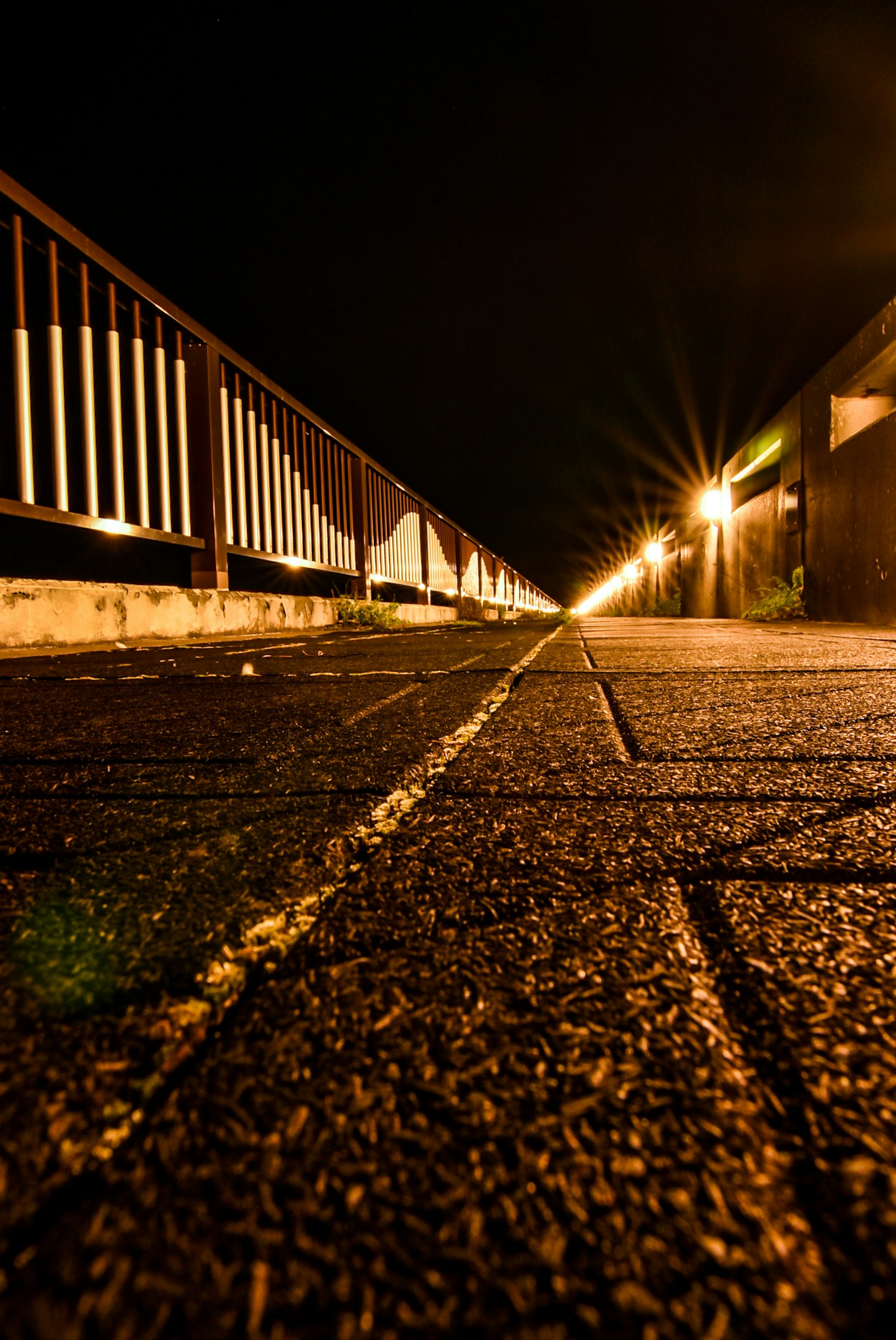 Photo of a walkway at night viewed from a low angle featuring illuminated railing and paved path