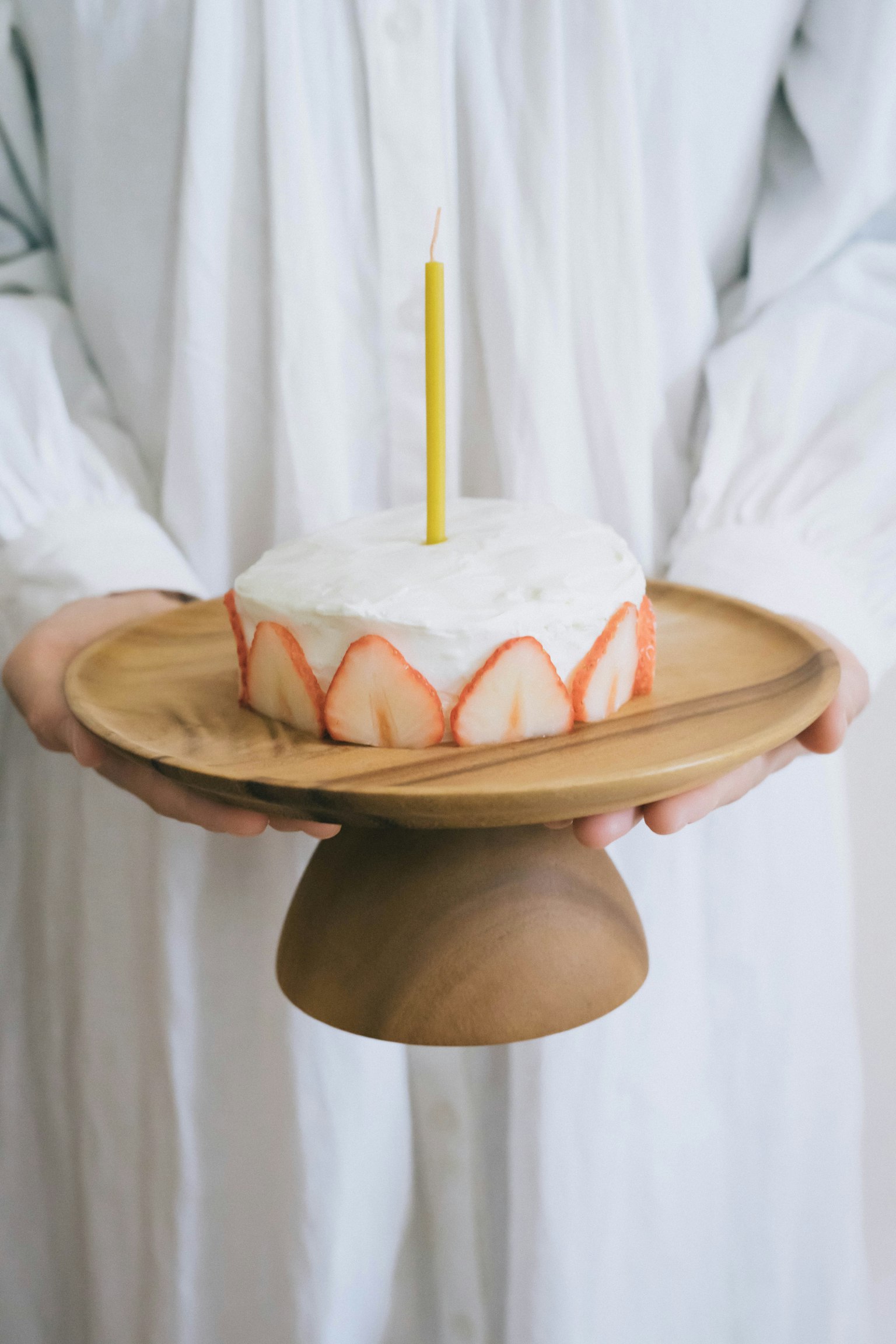 A person in a white dress holding a cake on a wooden stand with a yellow candle in the center