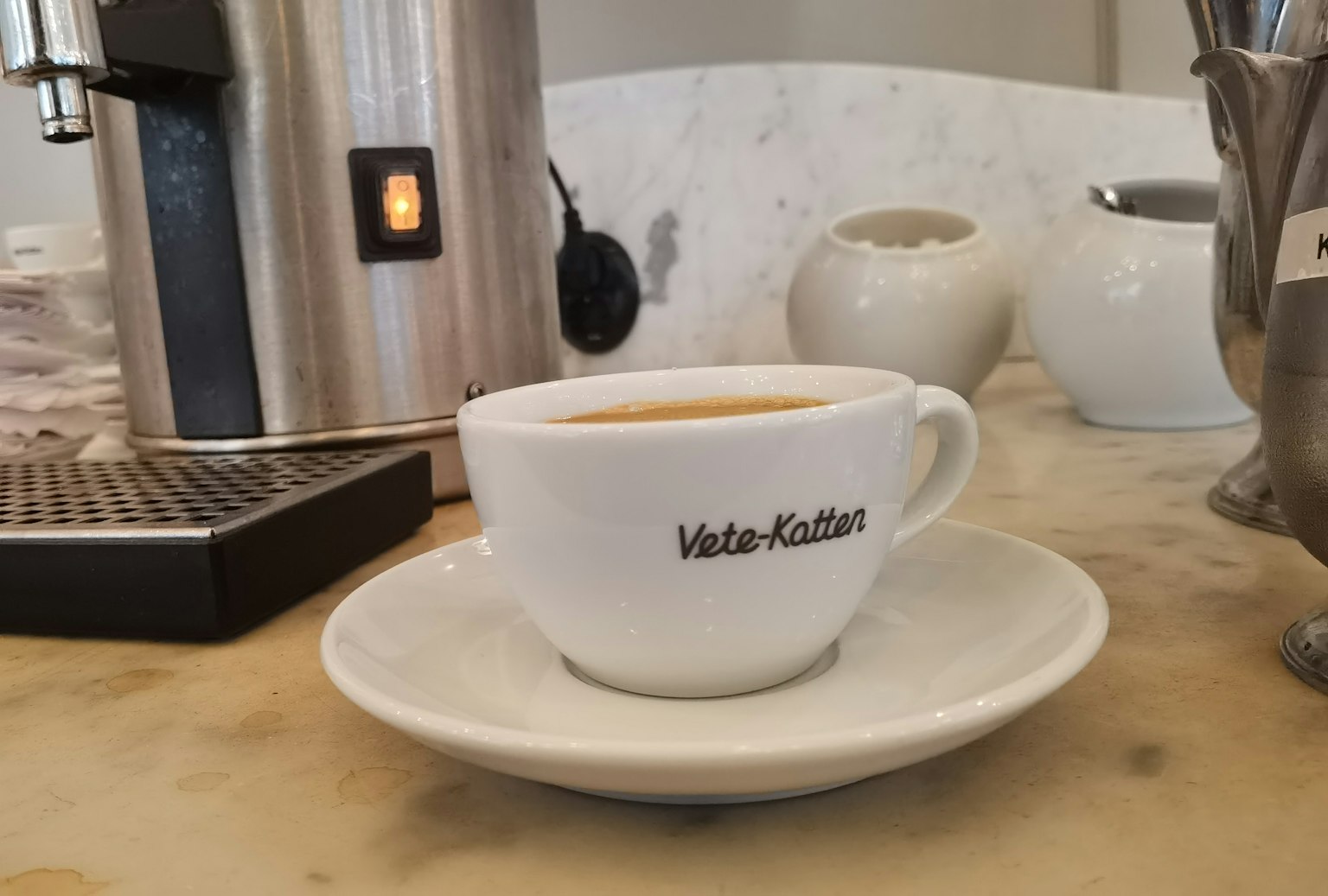 Cangkir kopi di atas piring di samping mesin espresso