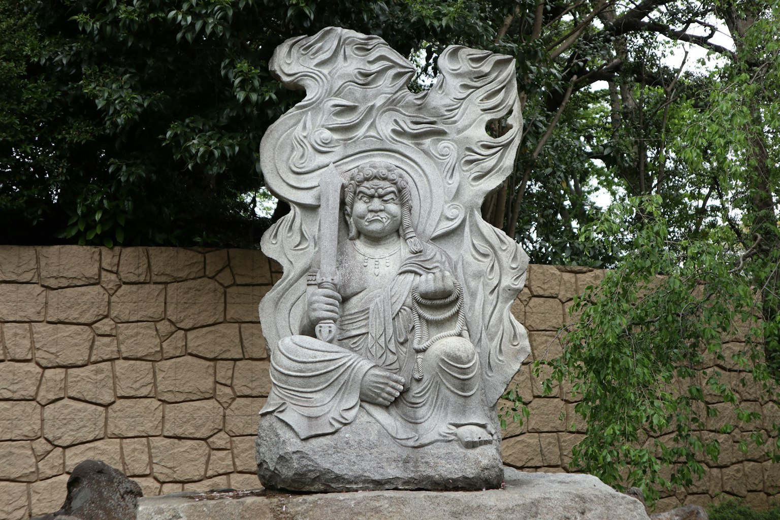 Sculpture en pierre d'une divinité assise entourée de verdure et d'un mur en pierre