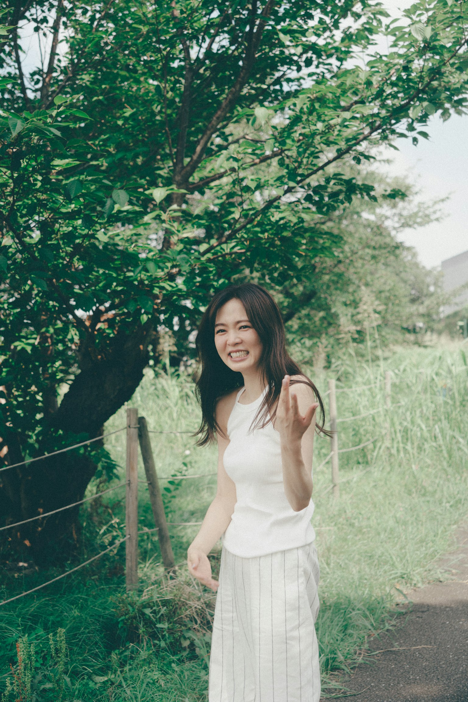 Một người phụ nữ mỉm cười và vẫy tay trước cây xanh mặc áo và váy trắng