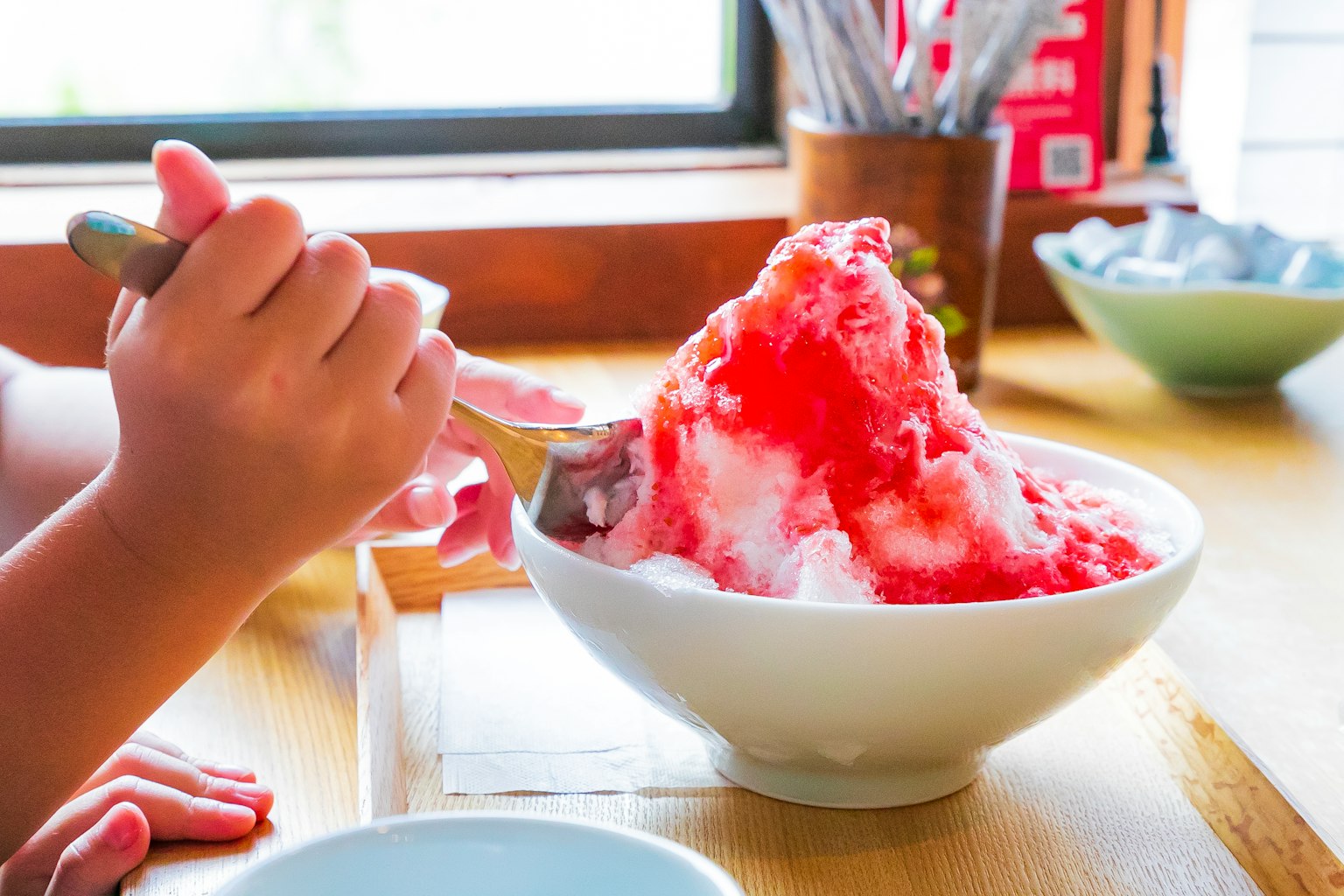 Hand holding a spoon with a bowl of shaved ice topped with red syrup