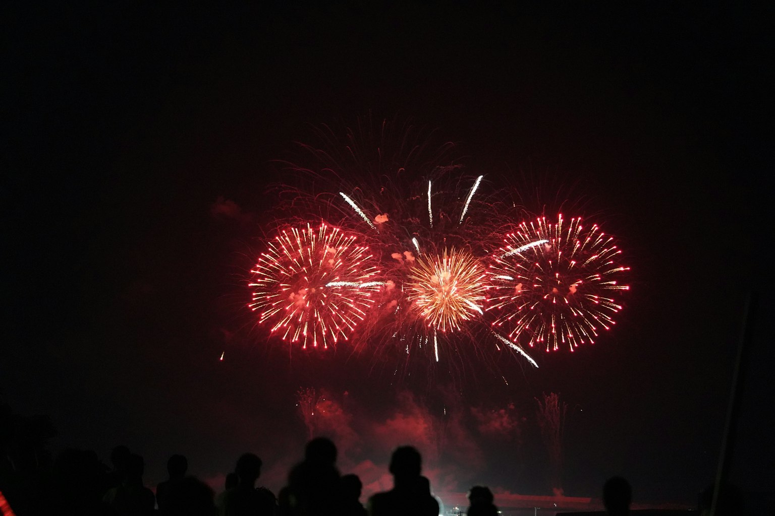 Tres fuegos artificiales rojos vibrantes estallando en el cielo nocturno con siluetas de personas en primer plano