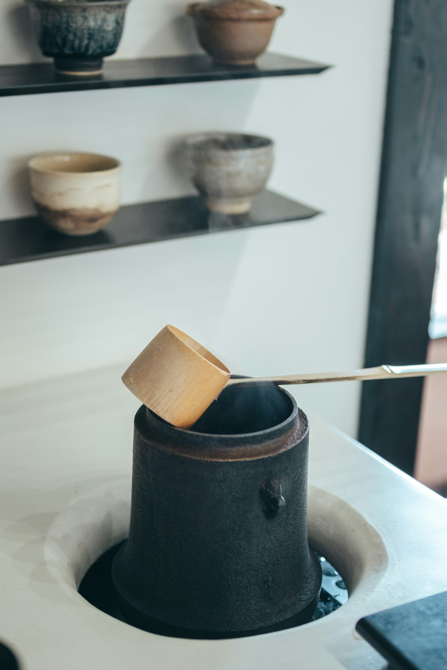 A wooden ladle resting on a black ceramic container with various pottery displayed on the wall shelf