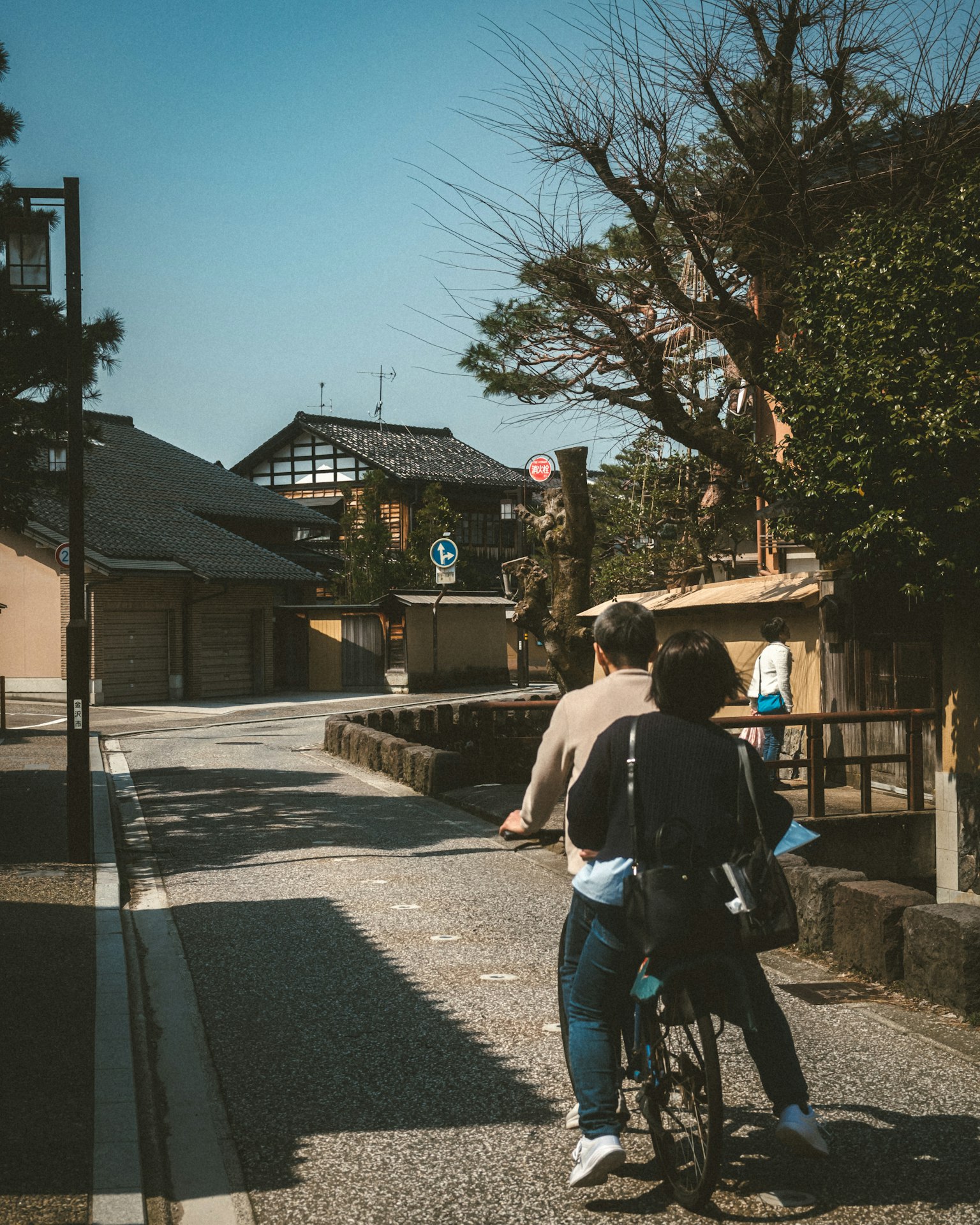 Un couple faisant du vélo sur une rue tranquille bordée de bâtiments japonais traditionnels et d'arbres verts