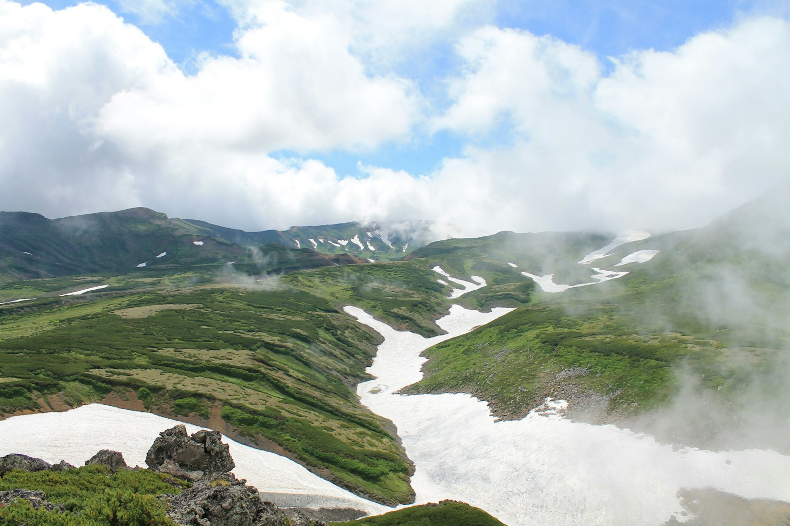 Lush green mountains with snow-covered valleys and drifting clouds in a blue sky