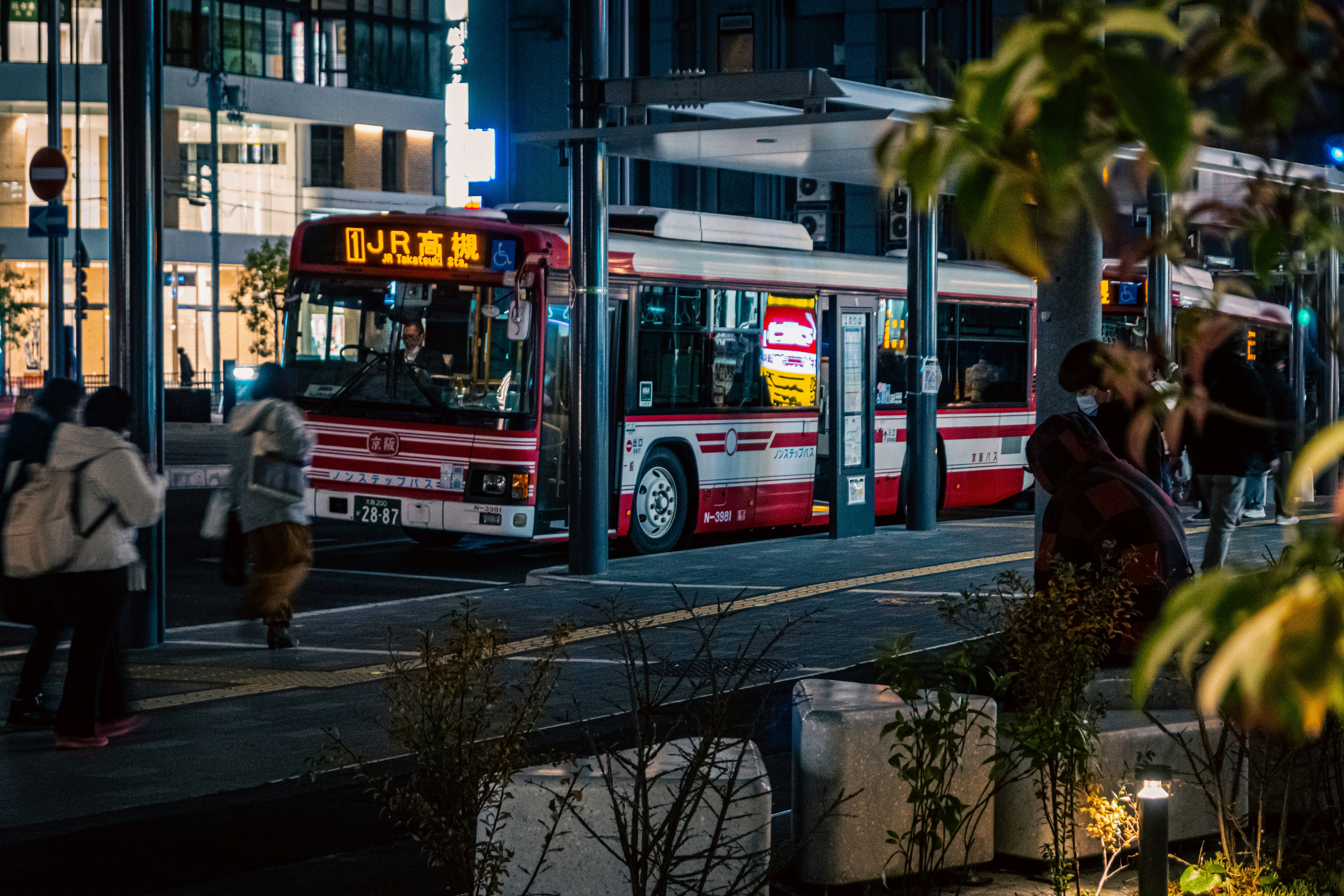 夜のバス停に停車中の赤と白のバスと周囲の人々