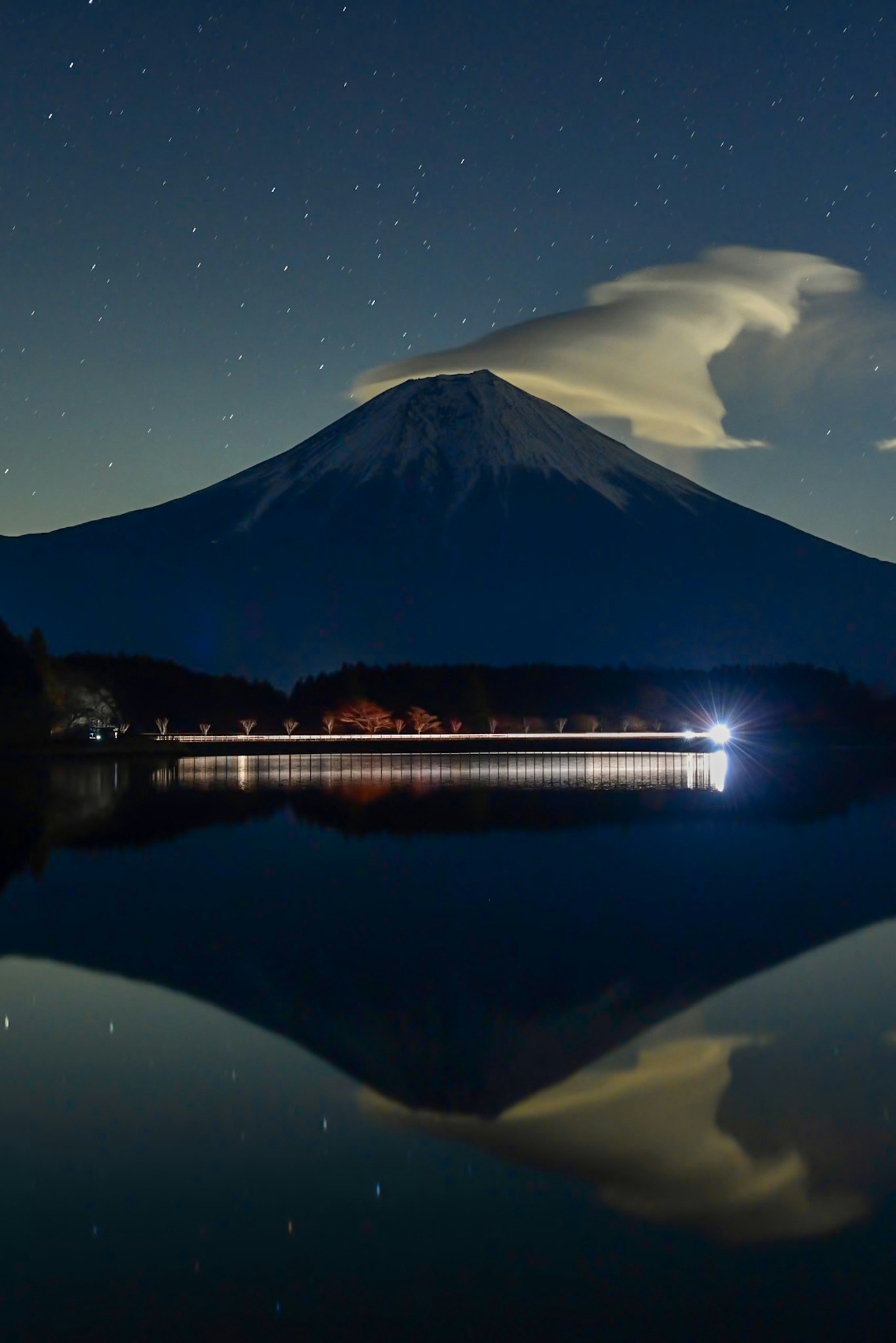 nachts reflektierter Berg im See mit Silhouette einer Brücke mit Lichtspuren