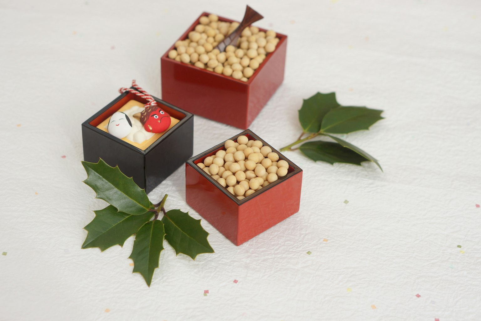 Three small decorative boxes in red and black colors filled with beans and topped with small ornaments surrounded by green leaves