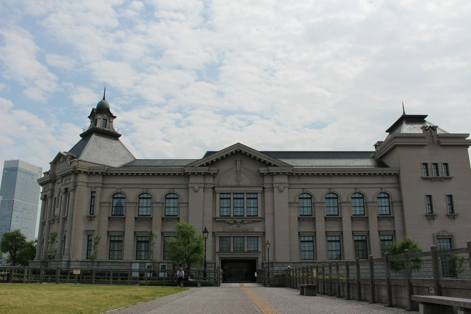 Façade d'un bâtiment historique avec ciel bleu et nuages en arrière-plan, mettant en valeur un aménagement paysager verdoyant
