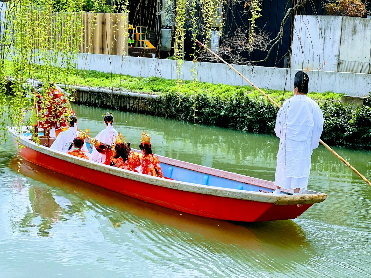 Sekelompok orang berwarna-warni dalam pakaian tradisional di perahu merah di saluran air yang tenang