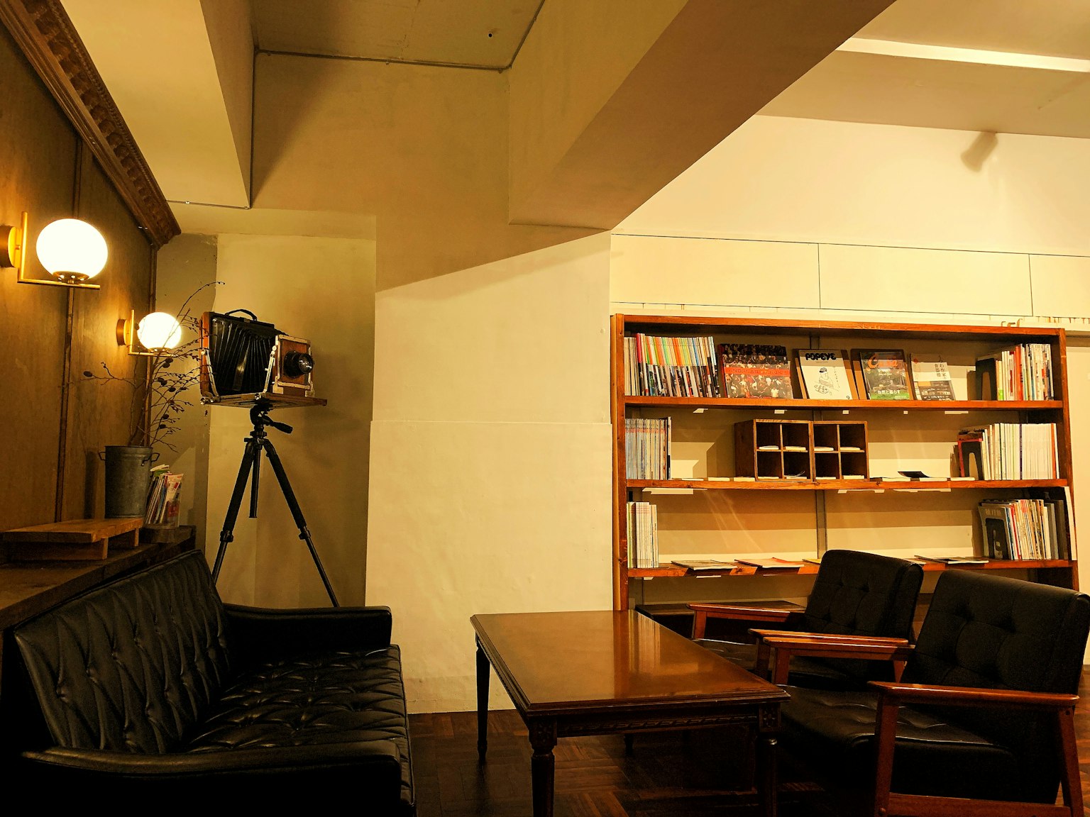Interior kafe yang nyaman dengan sofa hitam dan meja kayu rak buku penuh dengan buku kamera di tripod di latar belakang