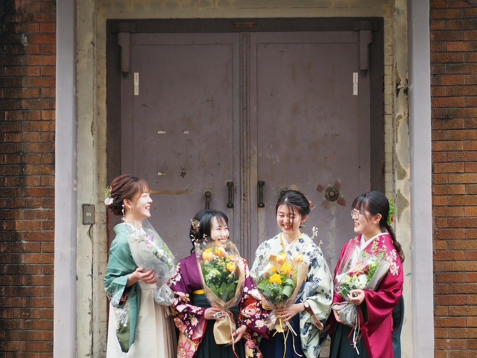 Four women in colorful kimonos holding flower bouquets smiling in front of a rustic door