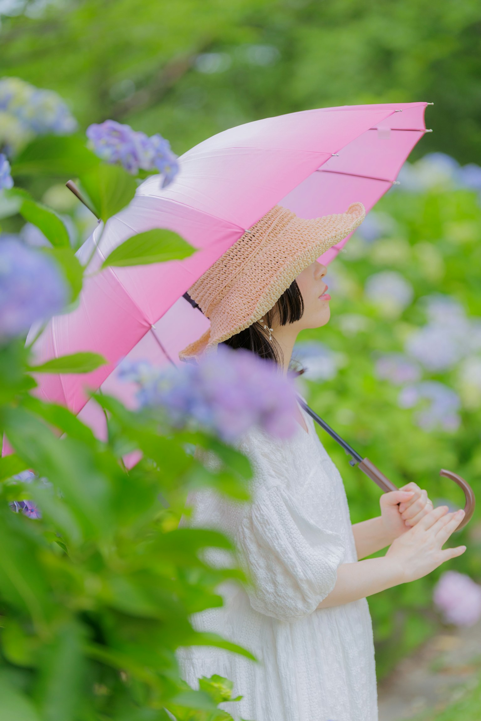 Una mujer con un vestido blanco sosteniendo un paraguas rosa rodeada de flores de hortensia