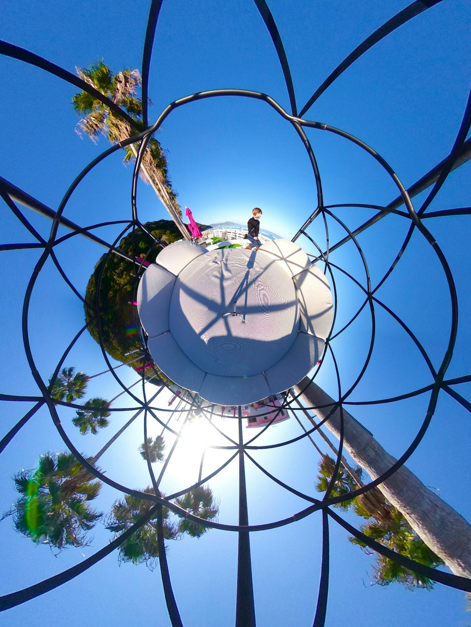 Cadre métallique entourant une structure de jeu circulaire sous un ciel bleu avec des palmiers