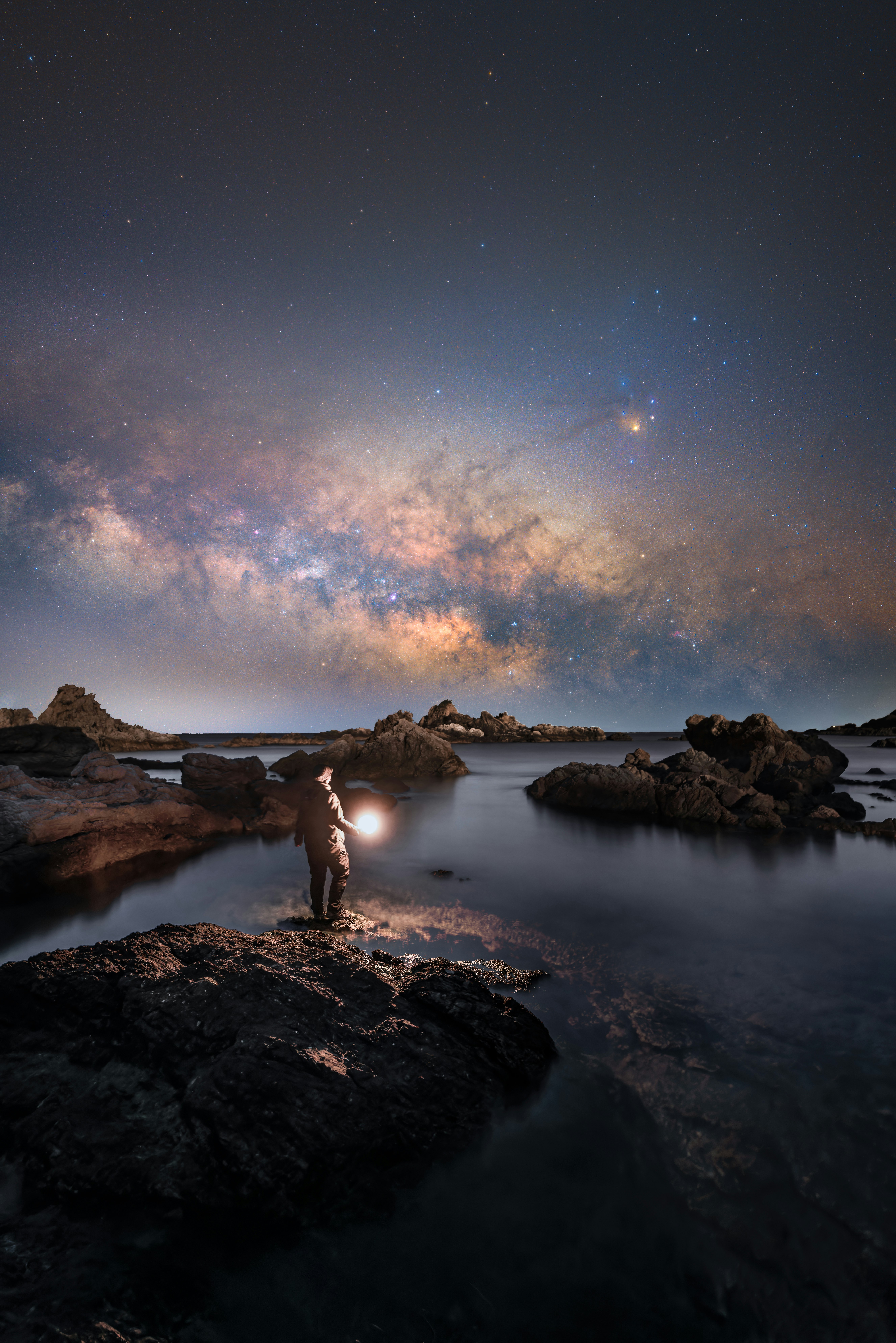 岩場の海岸で星空と天の川を見上げる人物の夜景