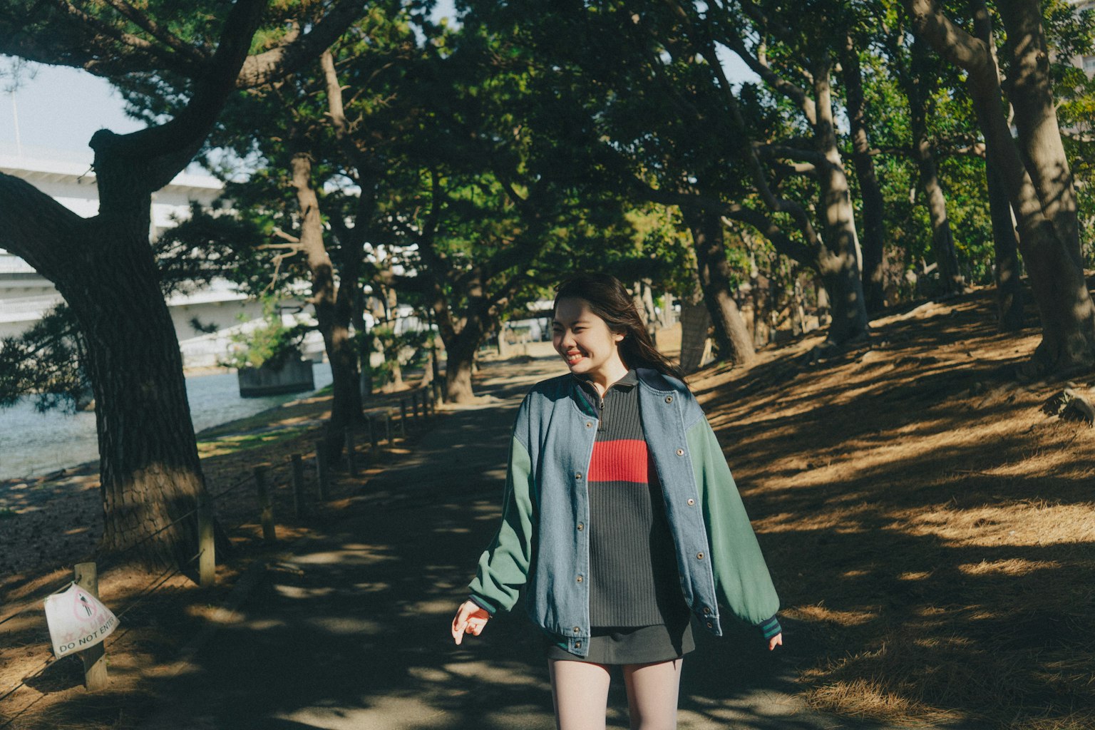 A woman walking among green trees wearing casual clothes and smiling brightly