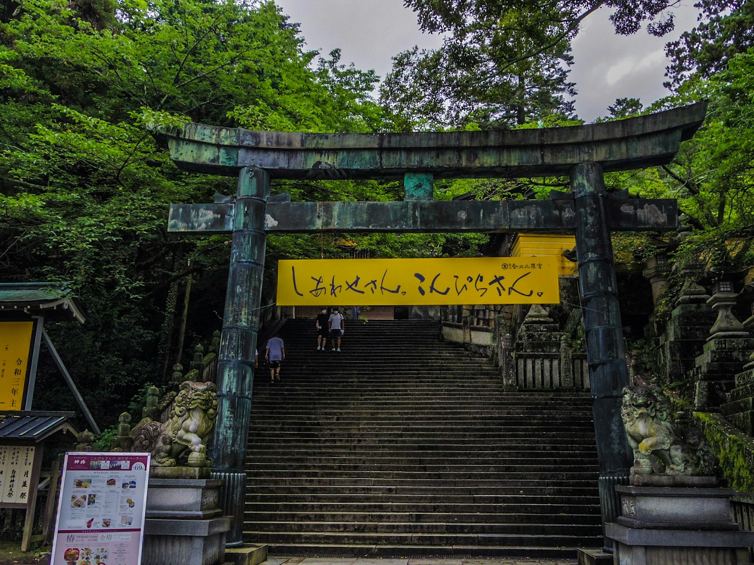 Un torii entouré de verdure avec des escaliers et un panneau jaune bien visible