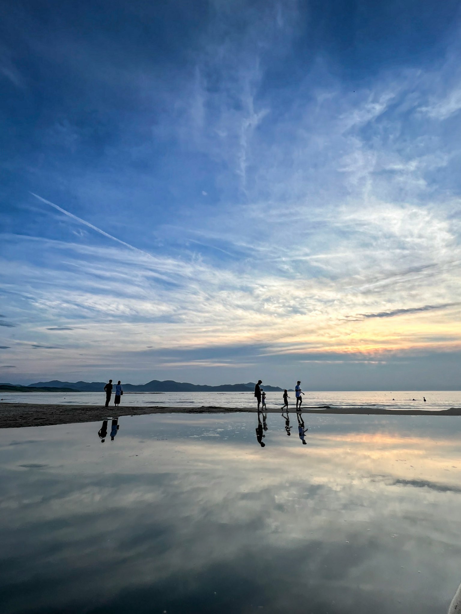 Silhouette di persone in riva al mare sotto un cielo blu con nuvole e riflessi sull'acqua