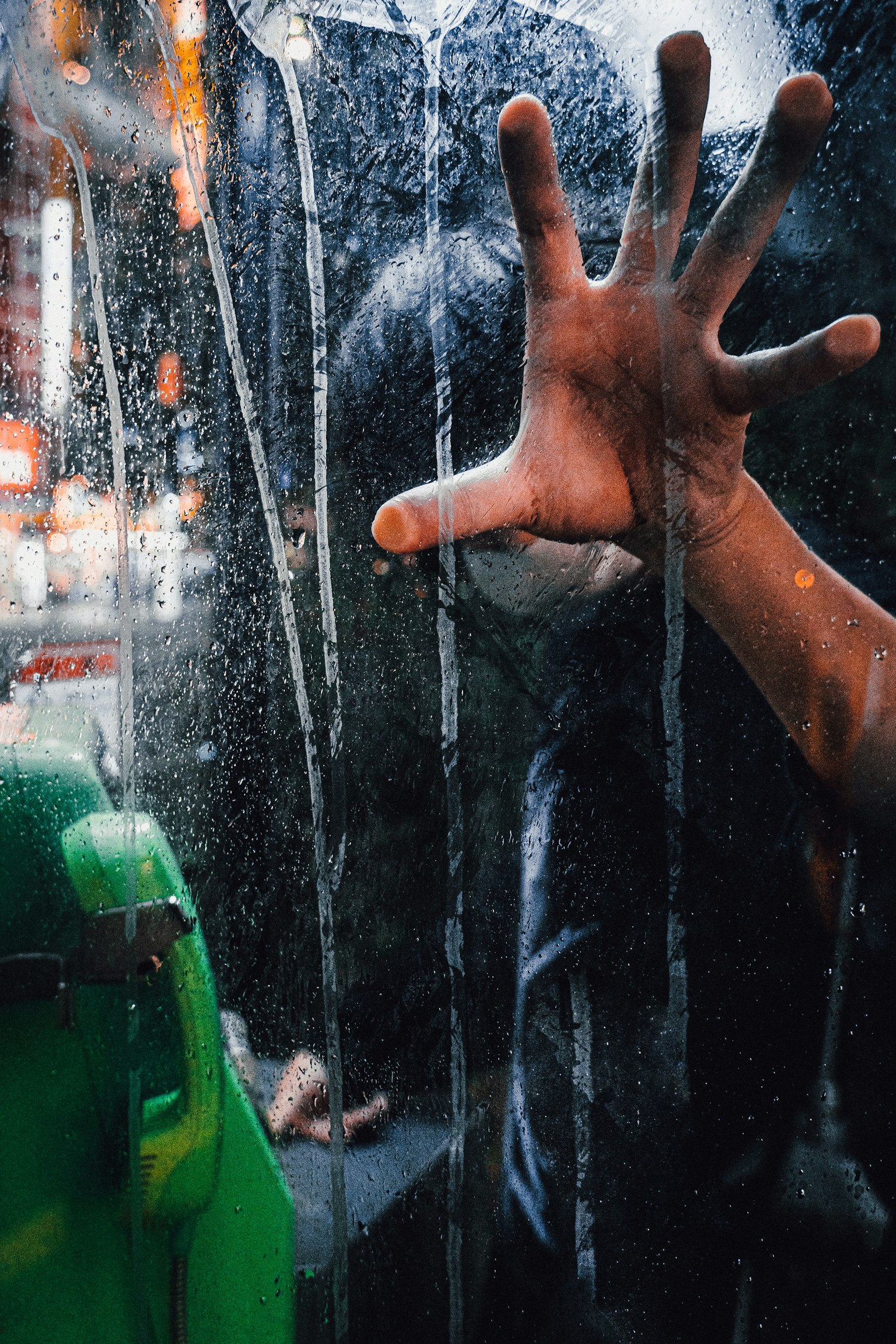 Silueta de una persona presionando la mano contra el vidrio bajo la lluvia con un objeto verde en primer plano