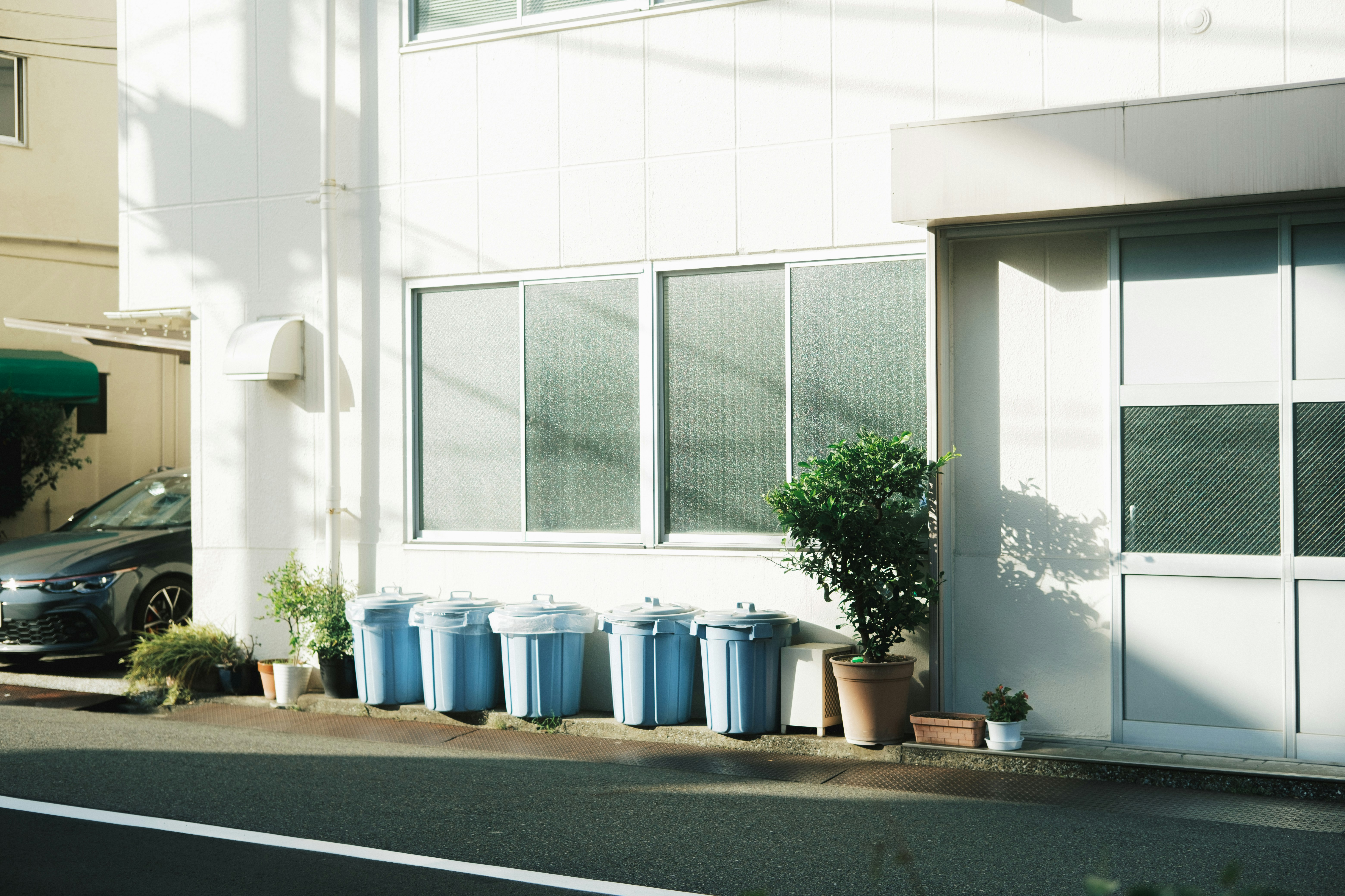 白い建物の軒先に並ぶ植木鉢と青いゴミ箱